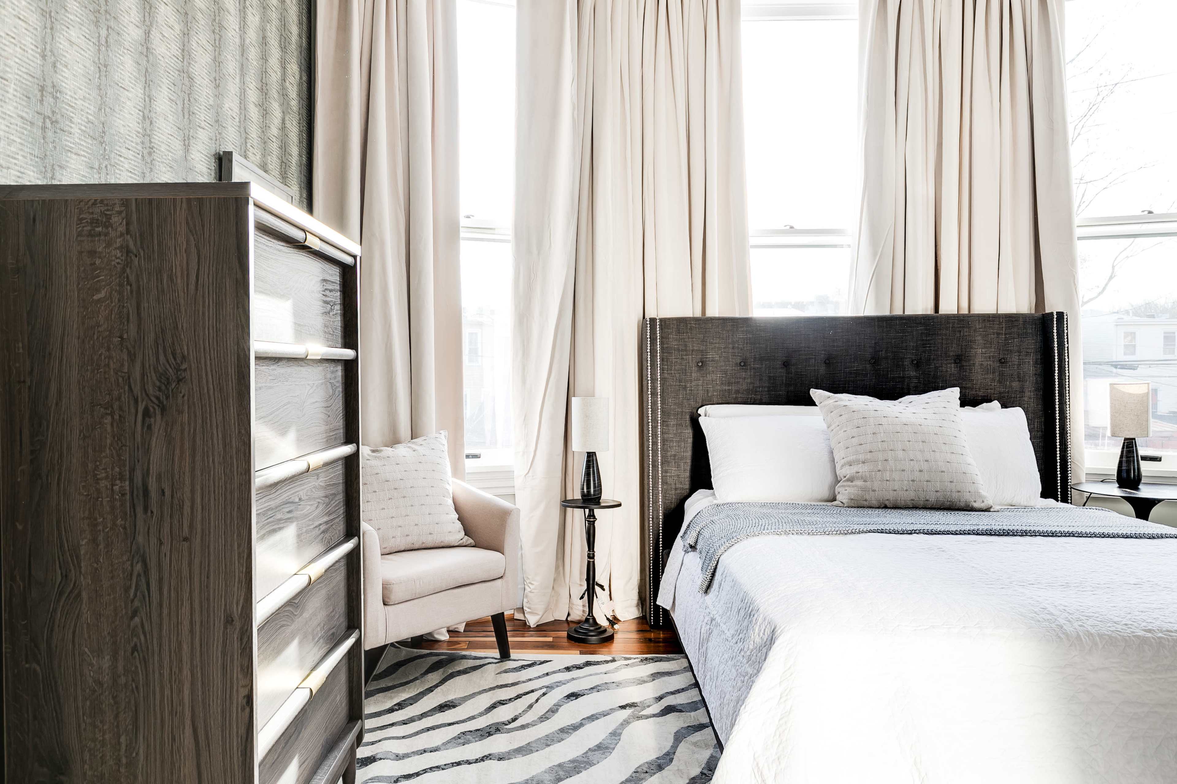 A softly lit bedroom featuring a bed with white linens, a patterned throw, a nightstand with a lamp, a gray dresser, and large windows with light-colored curtains.