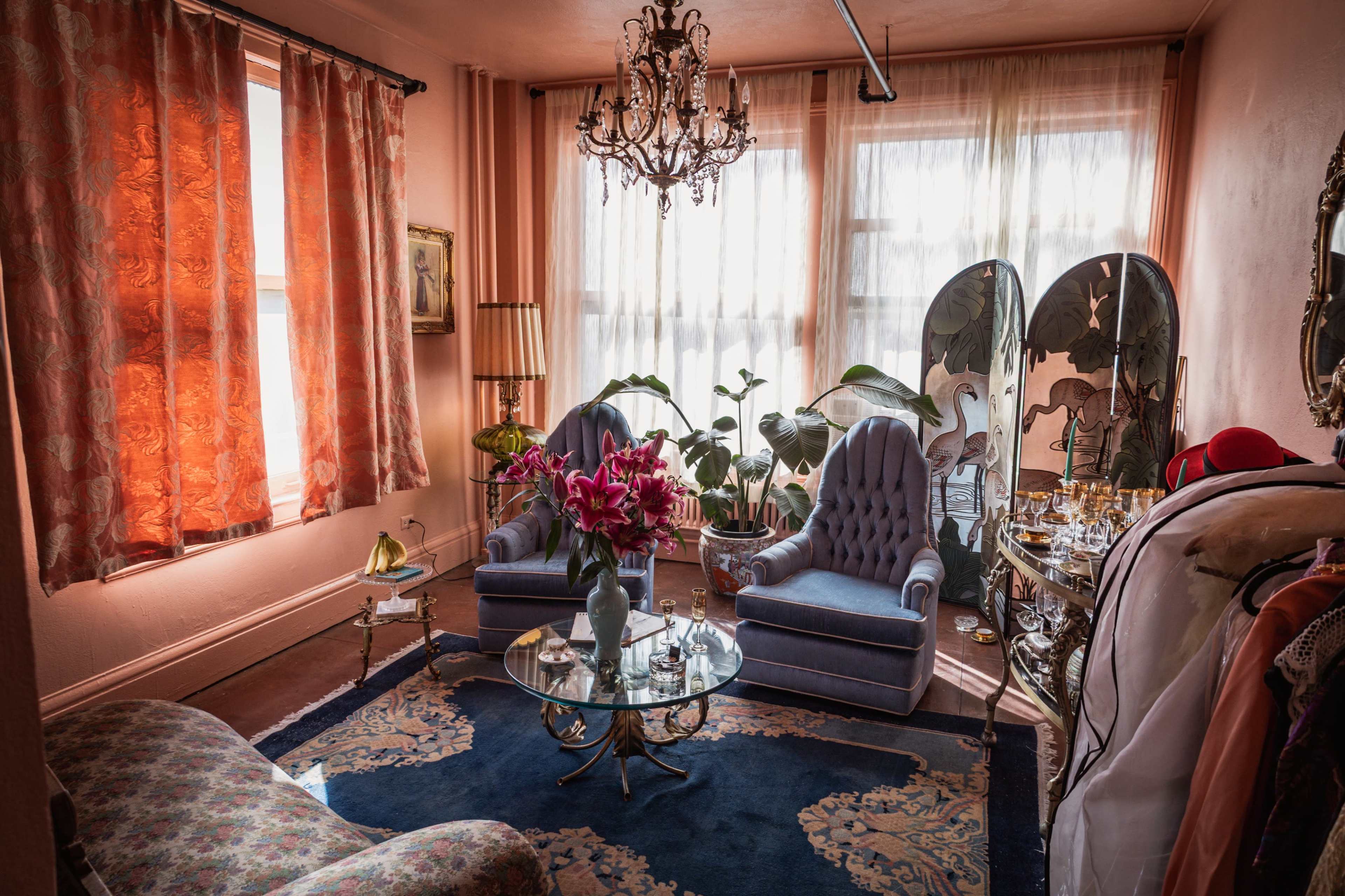 The image shows a cozy living room featuring two patterned chairs, a glass coffee table with a floral arrangement, and large windows draped with sheer curtains.