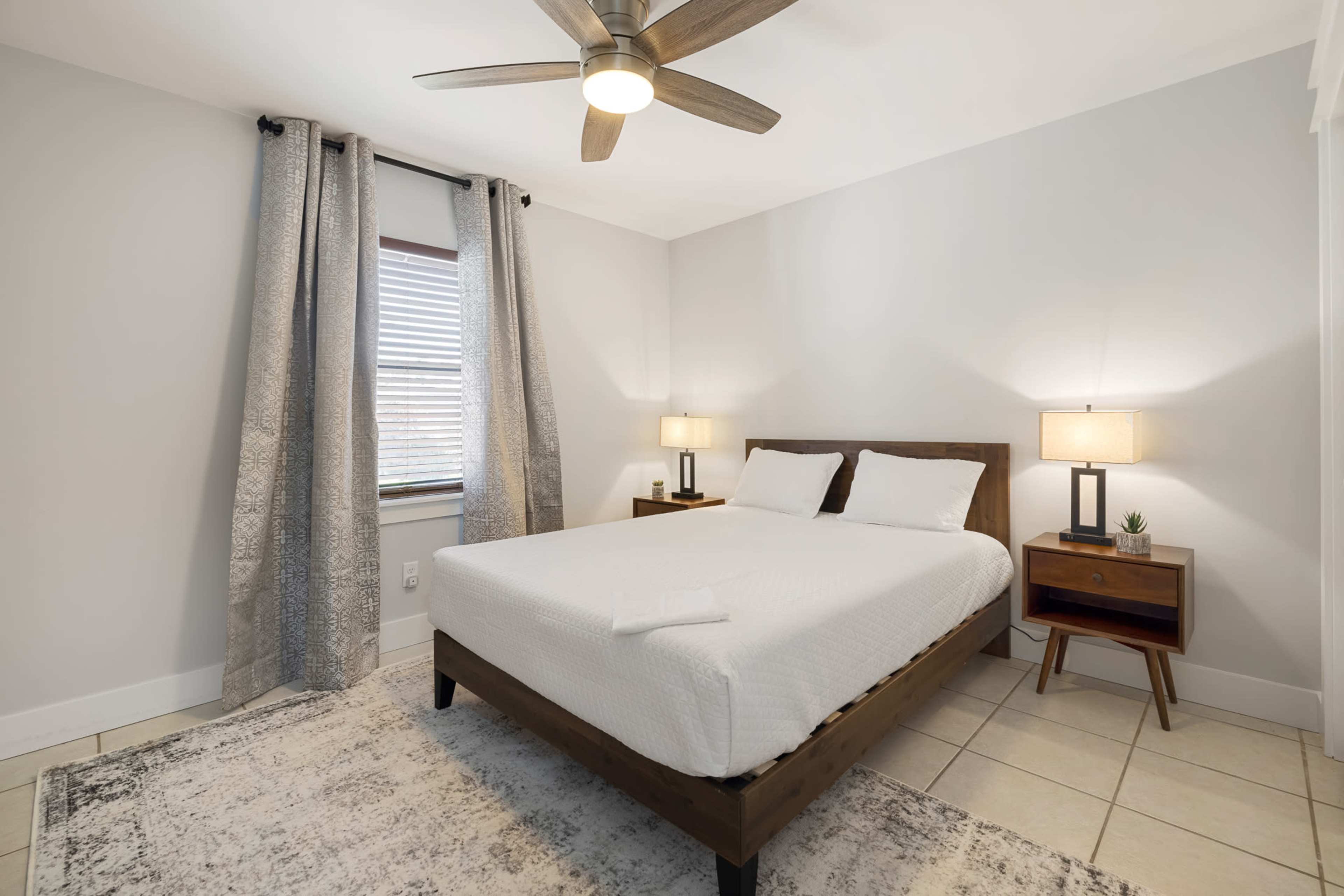 A tidy bedroom with a queen-sized bed, a nightstand with a lamp, and large windows covered by grey curtains.