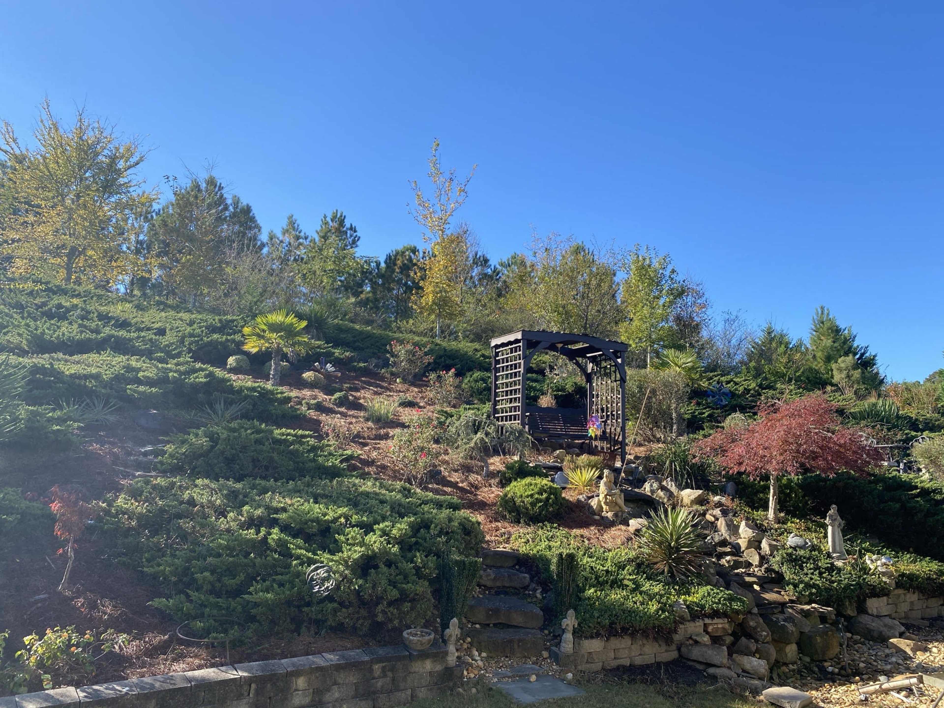 A wooden gazebo surrounded by landscaped greenery and stone steps is set against a clear blue sky.
