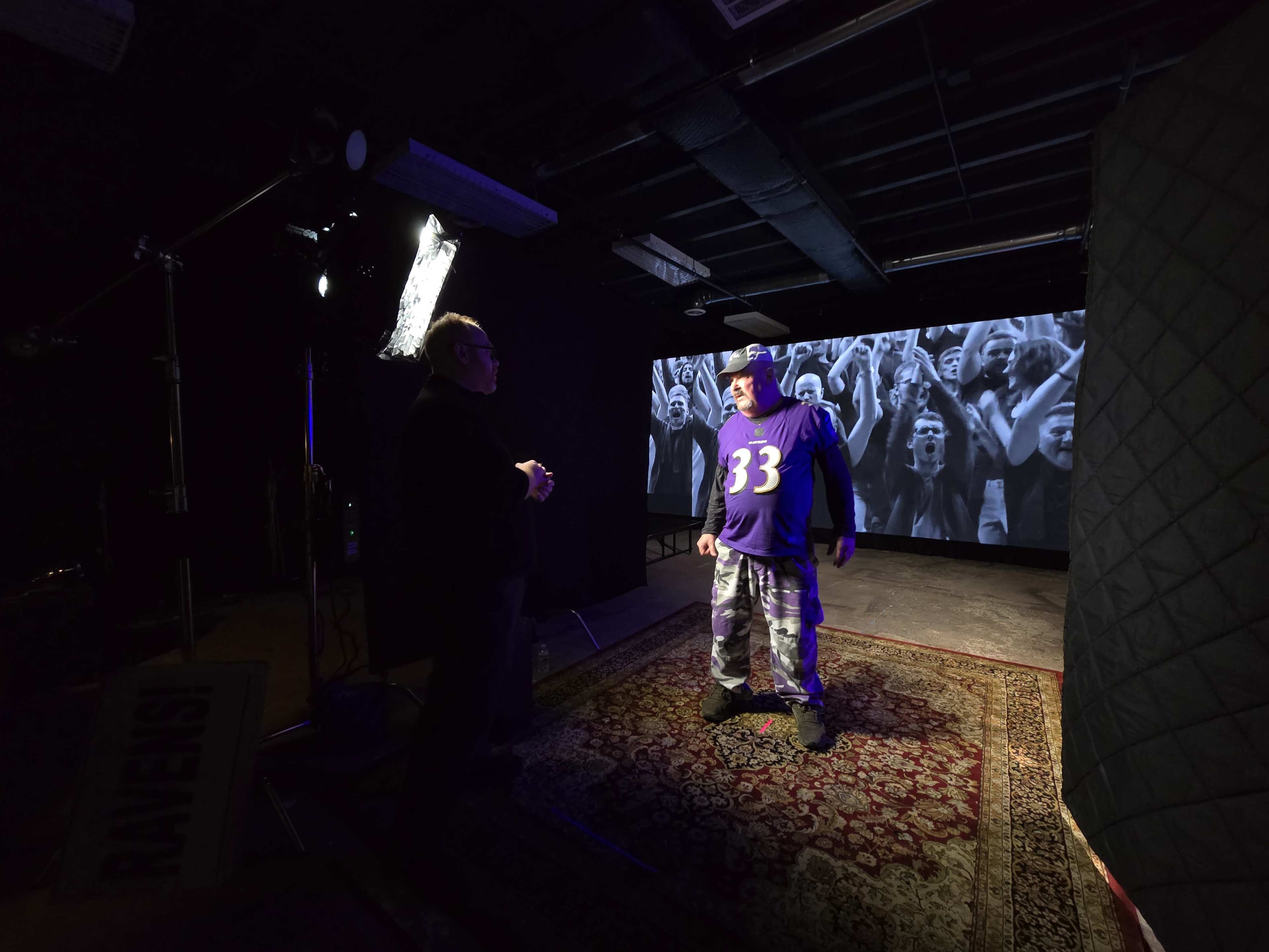 A man wearing a purple jersey and camouflage pants stands in front of a backdrop with cheering fans while conversing with another person.