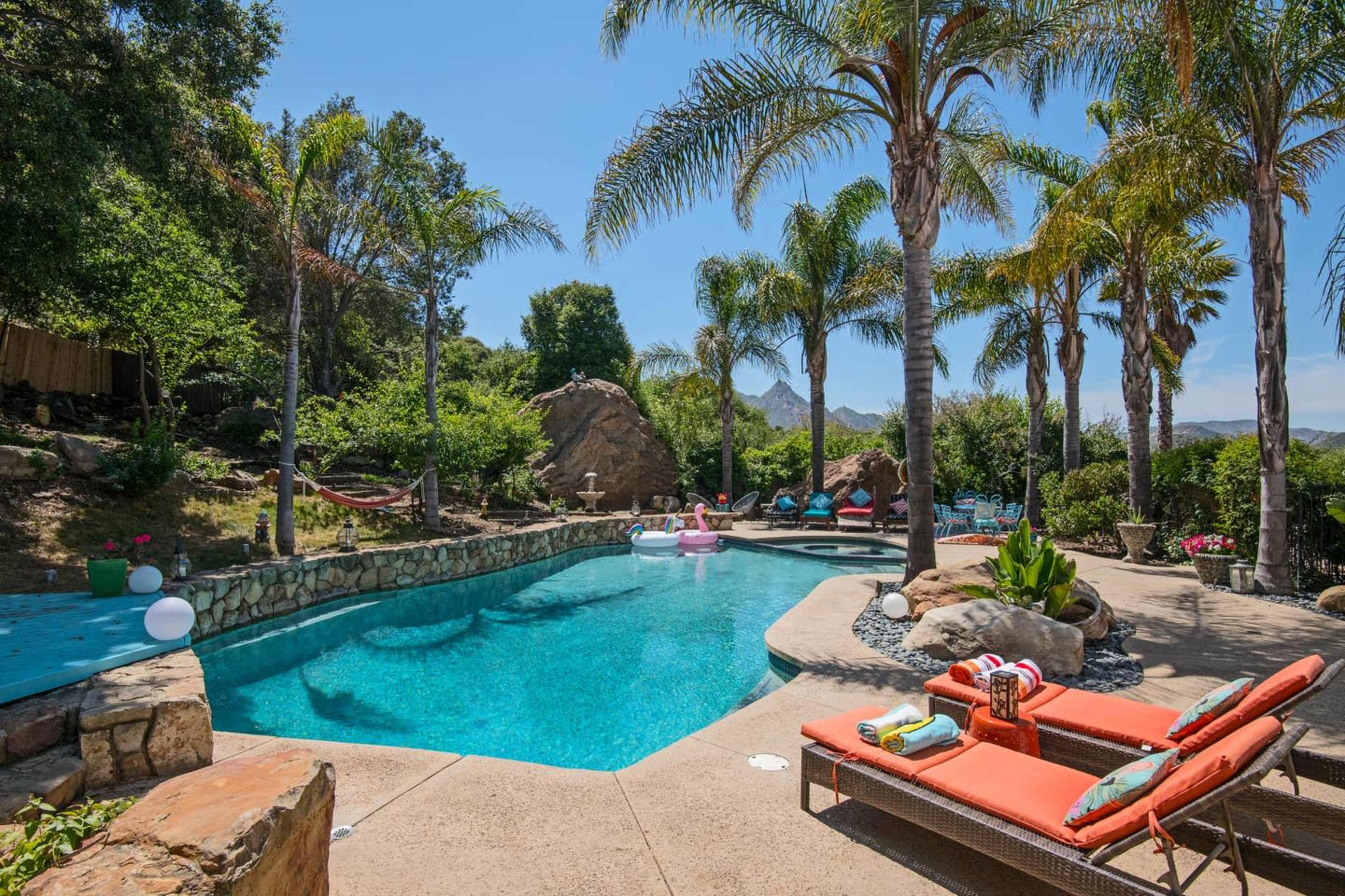 The image shows a backyard pool area surrounded by palm trees and lush greenery, featuring lounge chairs and decorative floats in the water.