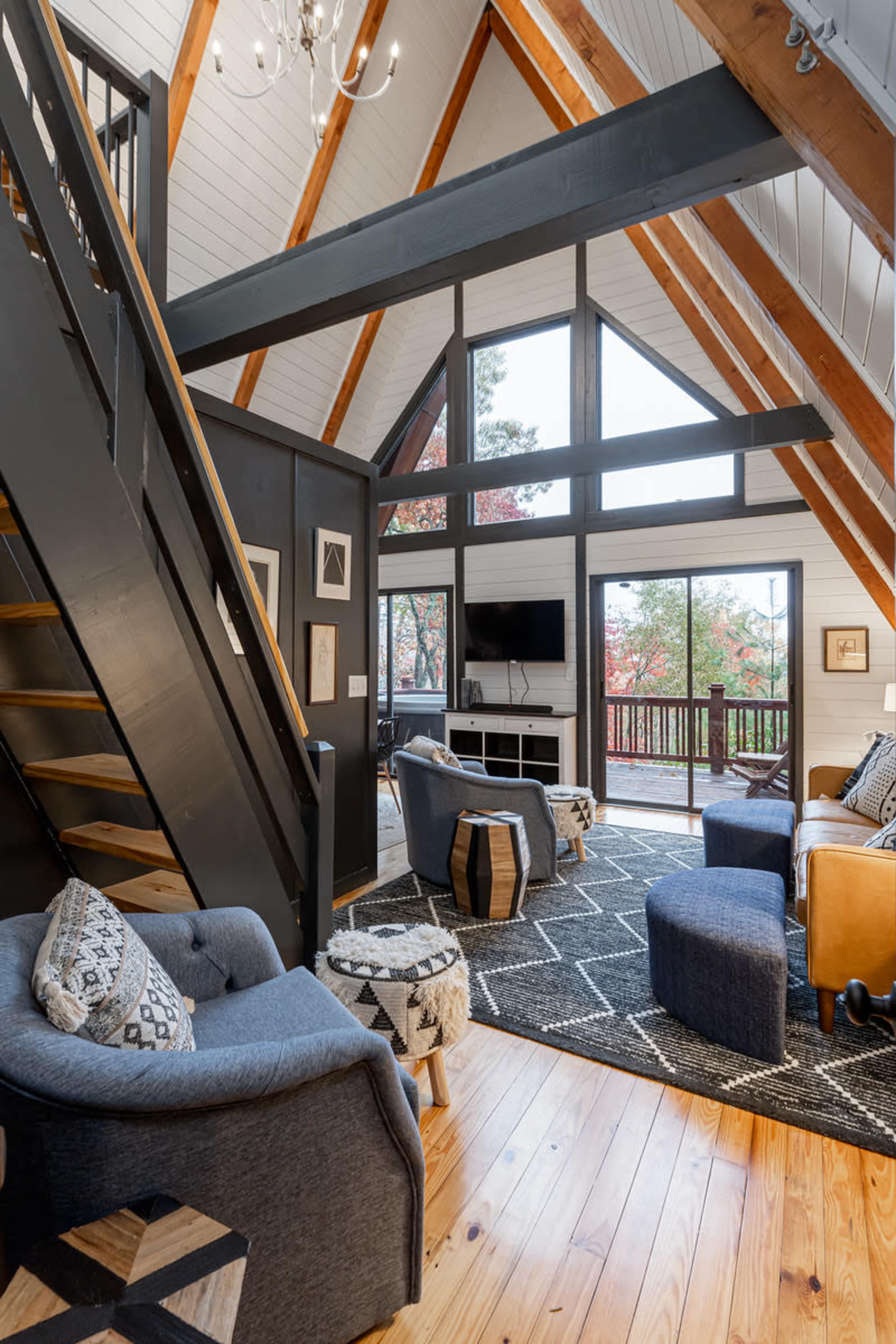 The image shows a spacious living room with wood beams, a staircase, and large windows offering a view of trees outside.