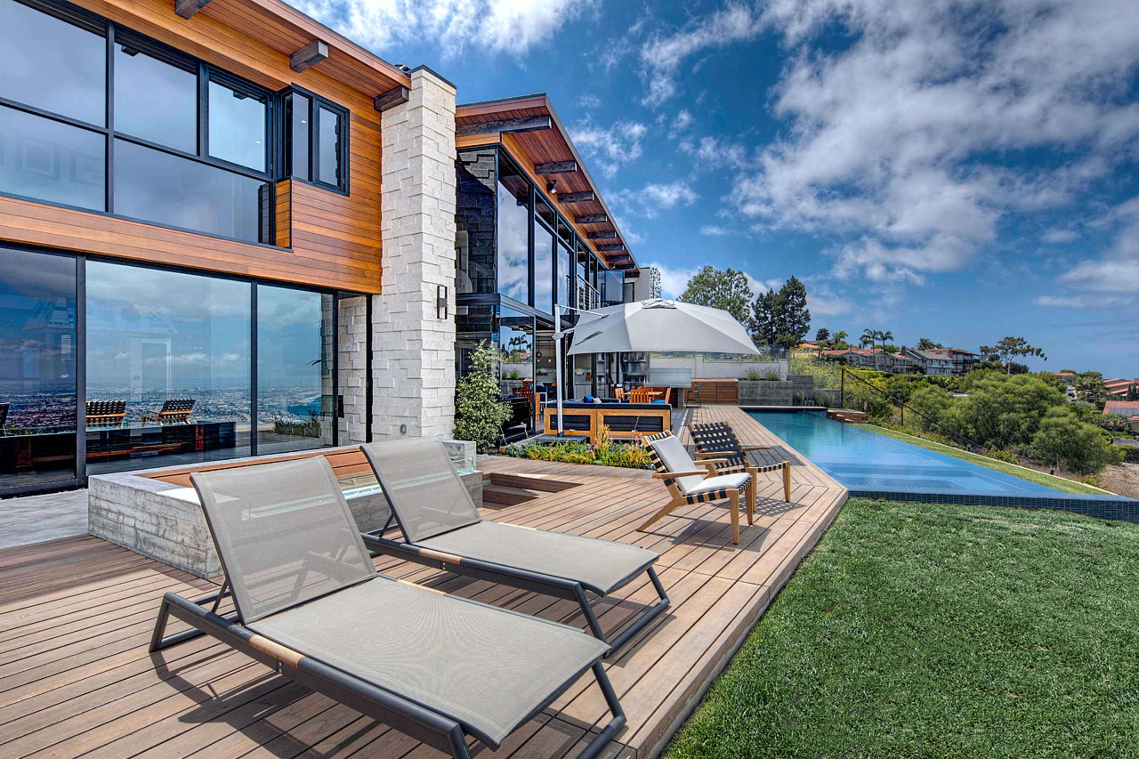 A modern home features a deck with lounge chairs beside an infinity pool overlooking a hillside view.