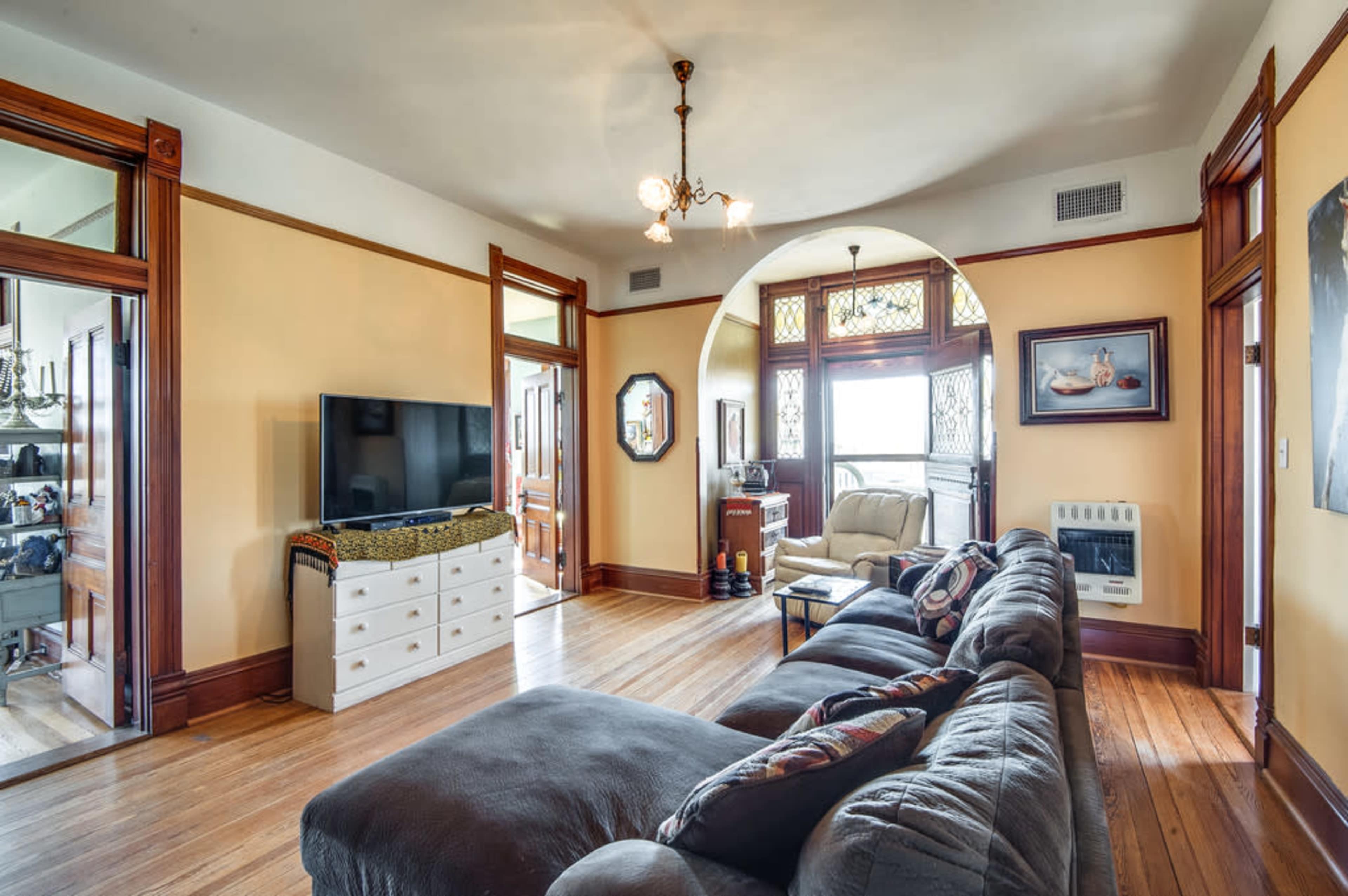 A living room with a gray sectional sofa, a television on a white dresser, and large windows providing natural light, framed by wooden architectural details.