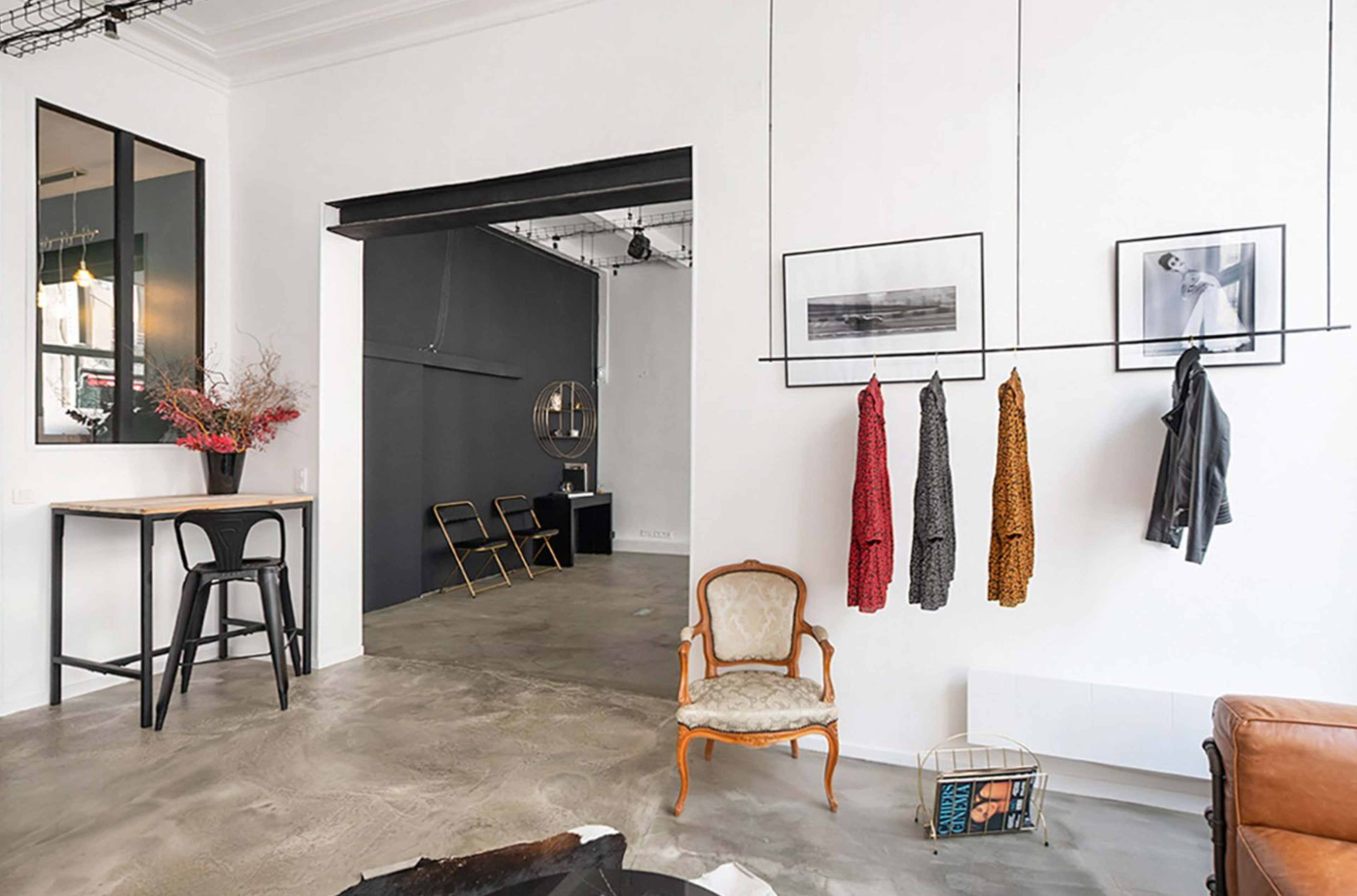 A minimalist interior featuring a small seating area with an antique chair, a table, and hanging clothing on display, along with a view of a darker room in the background.