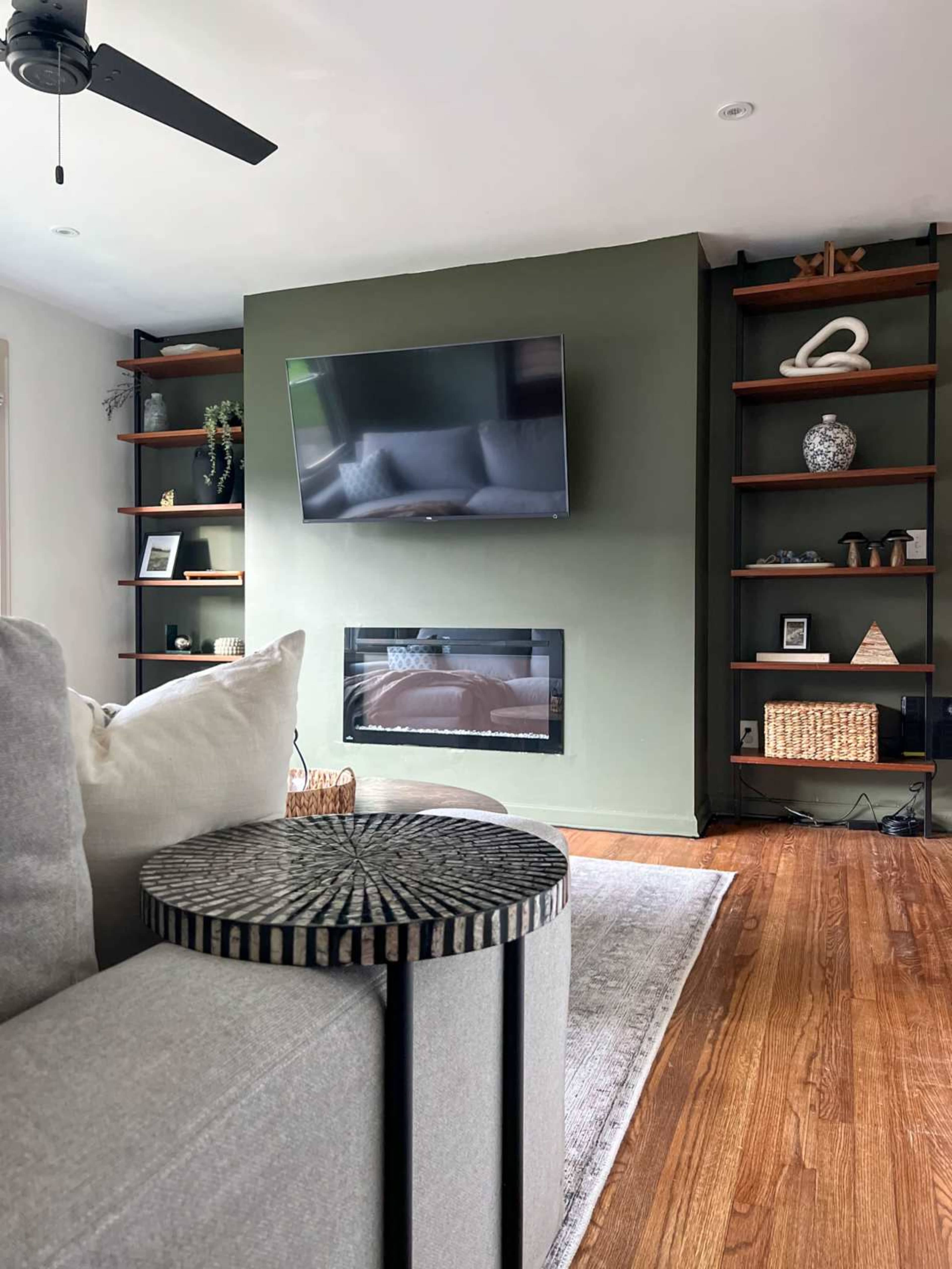 A cozy living room features a gray sectional sofa, a circular side table, a mounted television above a modern fireplace, and dark wooden shelves displaying various decorative items.