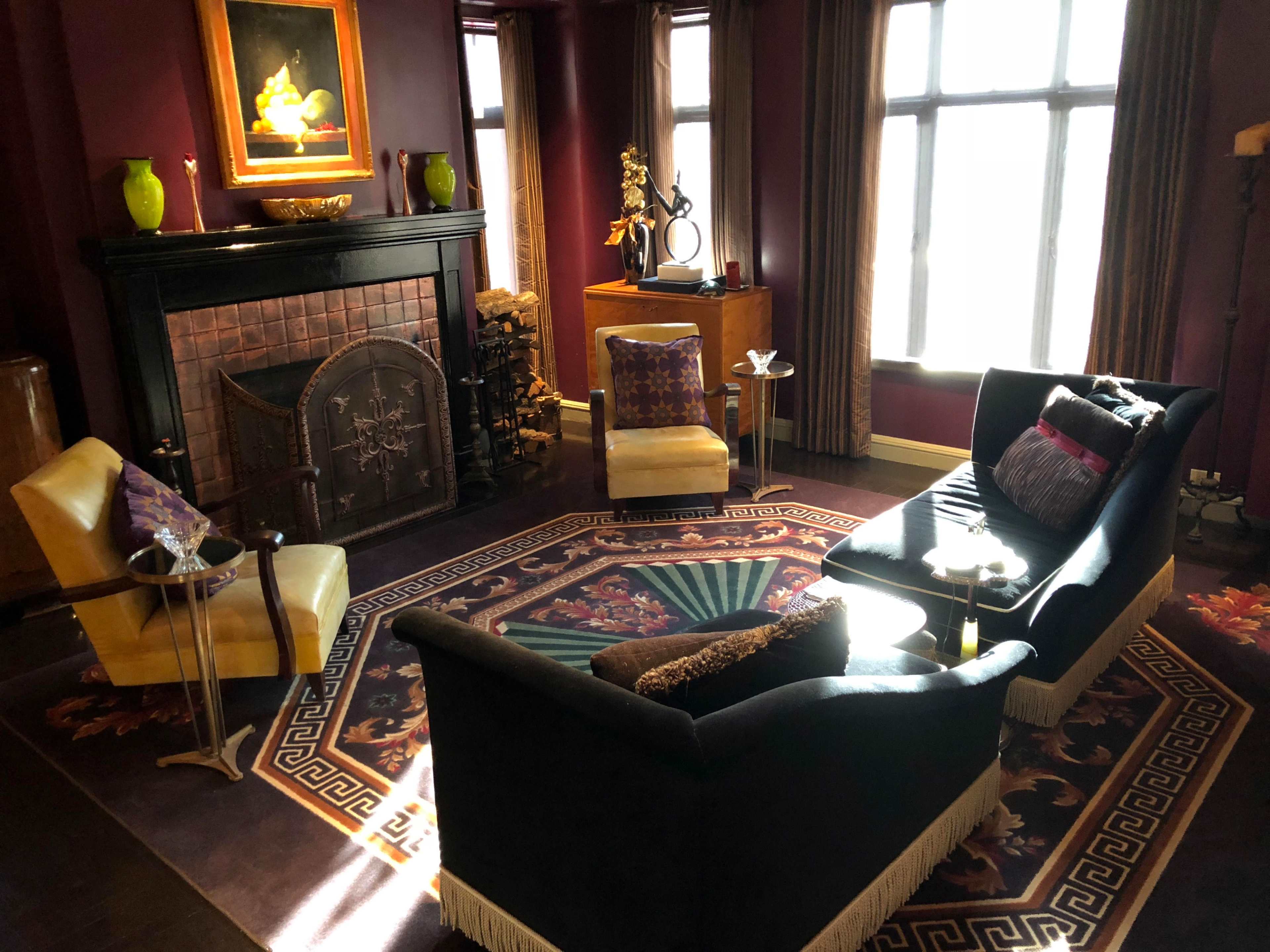 A sitting room features two armchairs and a sofa arranged around a decorative rug, with a fireplace and large windows allowing light to enter.