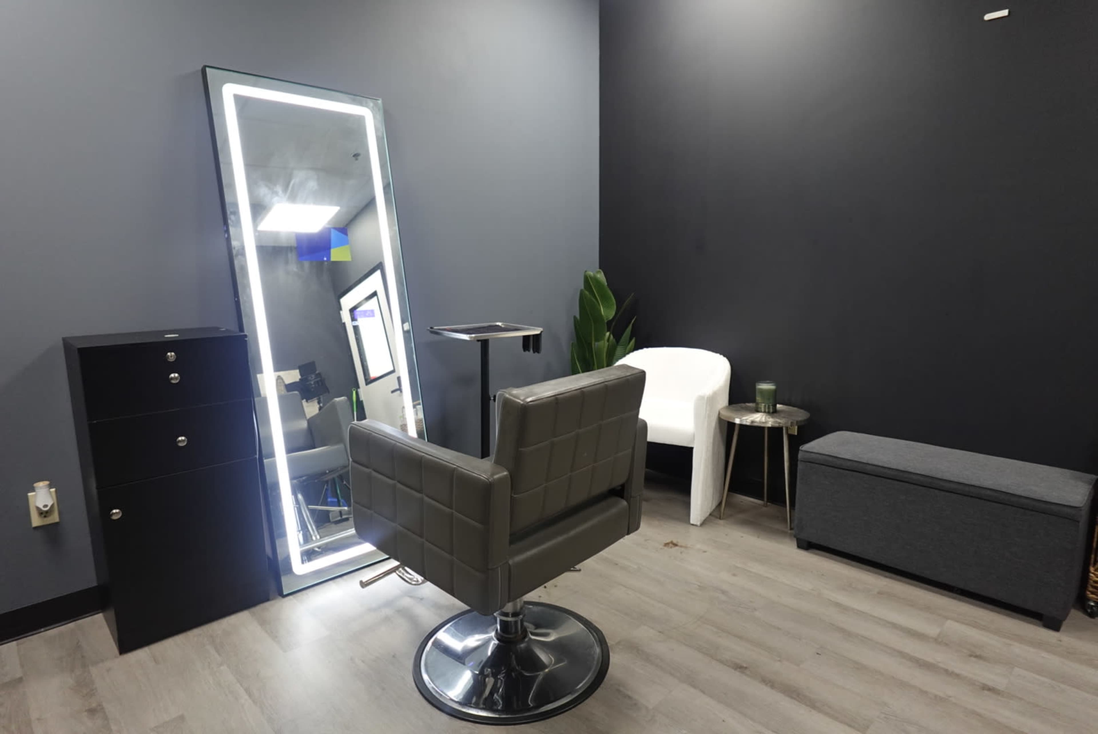 The image shows a minimalist salon interior featuring a gray chair, a large illuminated mirror, a small side table, and a storage cabinet against a dark wall.