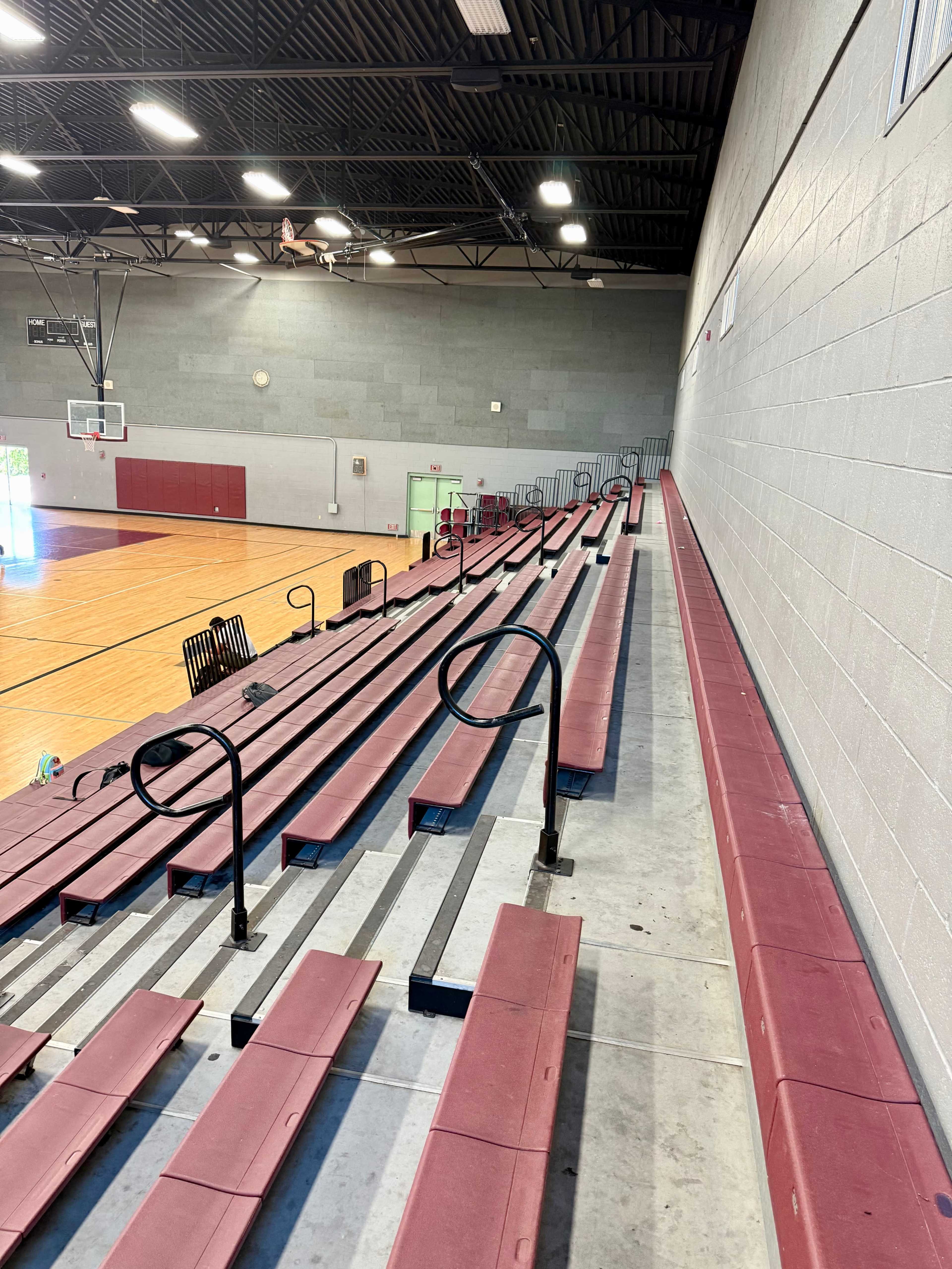 The image shows empty red and black bleachers in a gymnasium, with wooden basketball court flooring visible in the background.