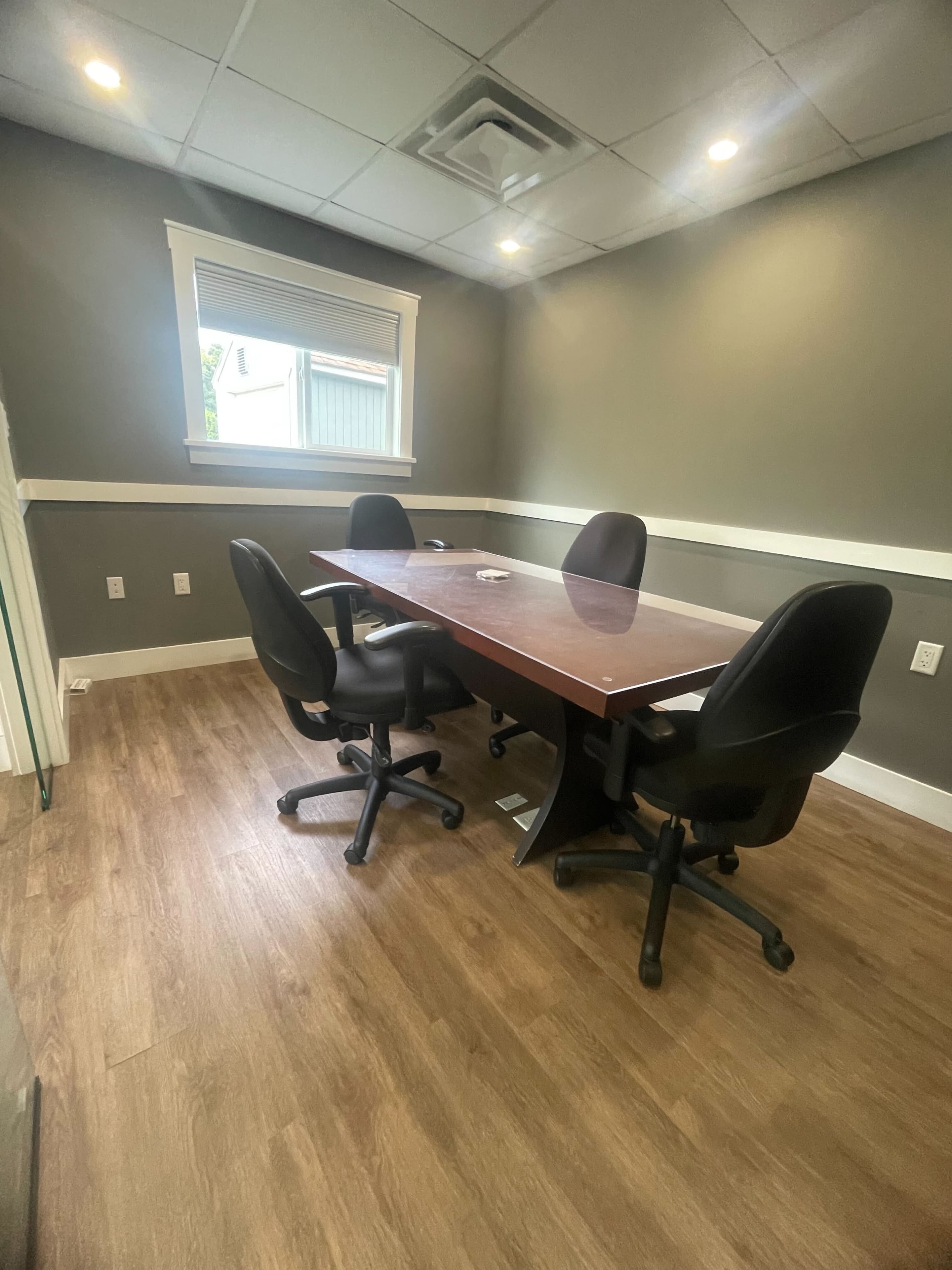 A conference room is depicted, featuring a rectangular wooden table surrounded by four black rolling chairs.