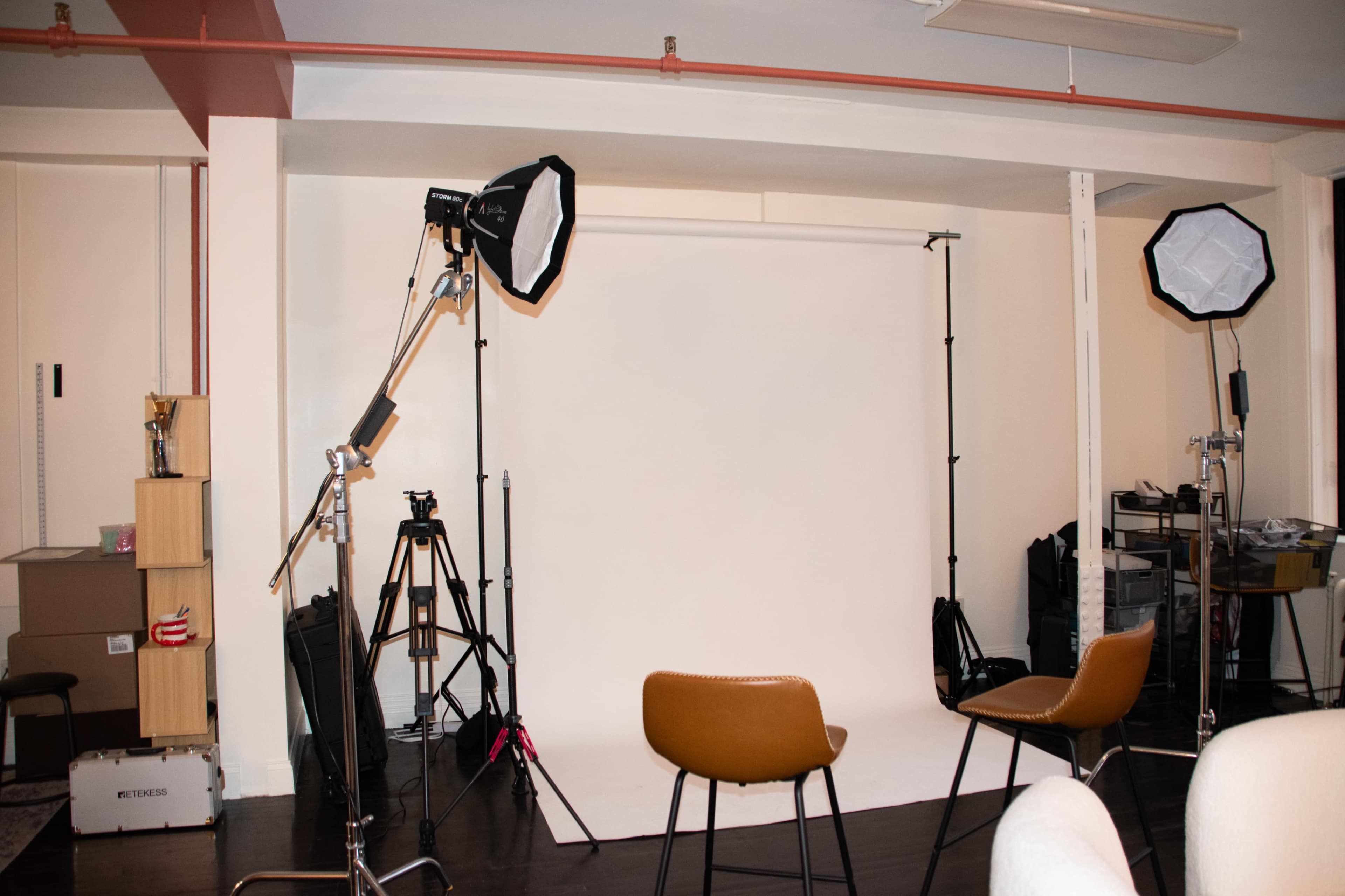 The image shows a photography studio setup with two softbox lights, a plain white backdrop, and two brown chairs positioned in front of it.