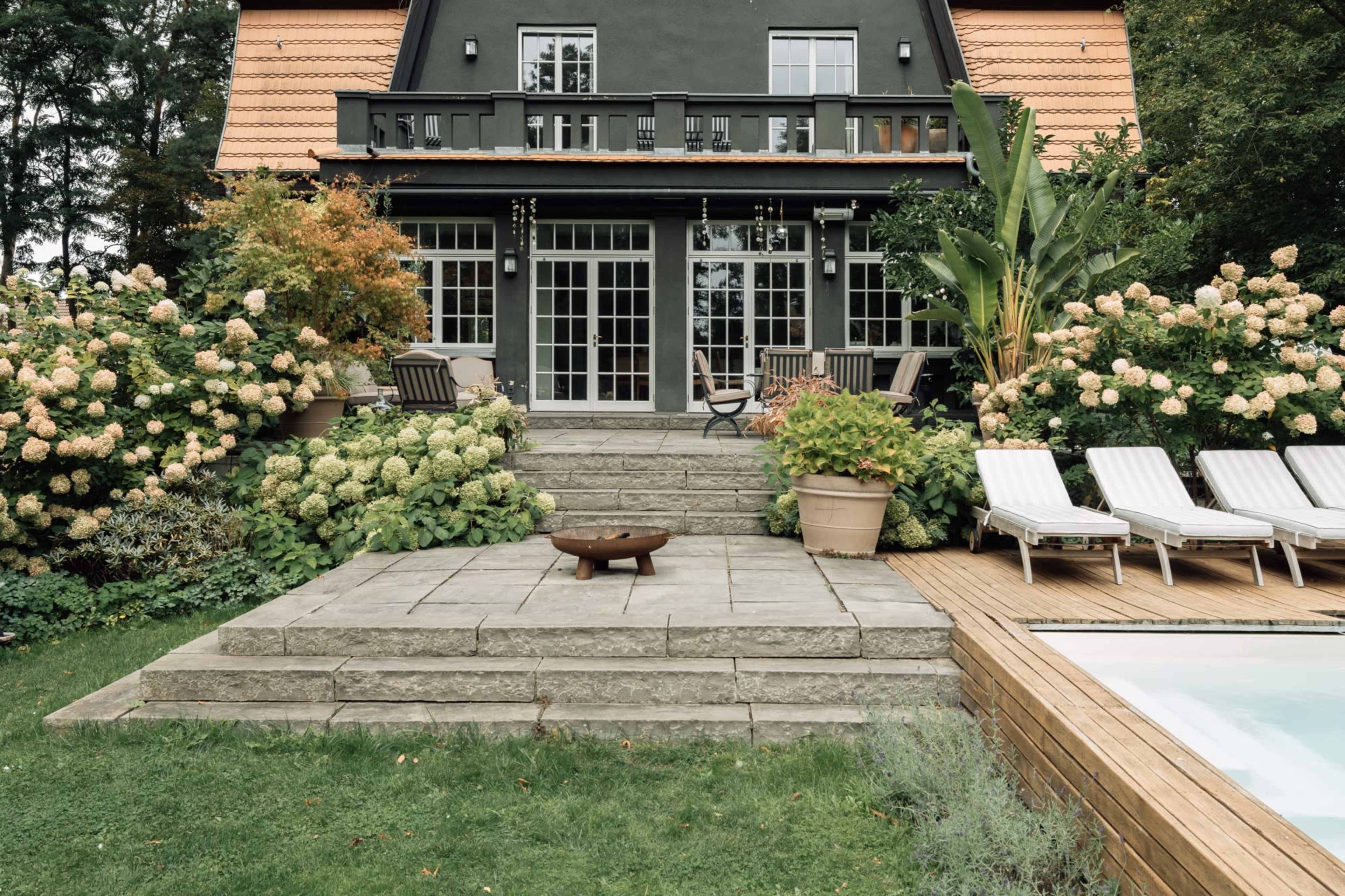A well-landscaped backyard features a gray stone patio with a fire pit, surrounded by lush greenery and flowering hydrangeas, adjacent to a swimming pool and a two-story house with a black exterior and orange roof.