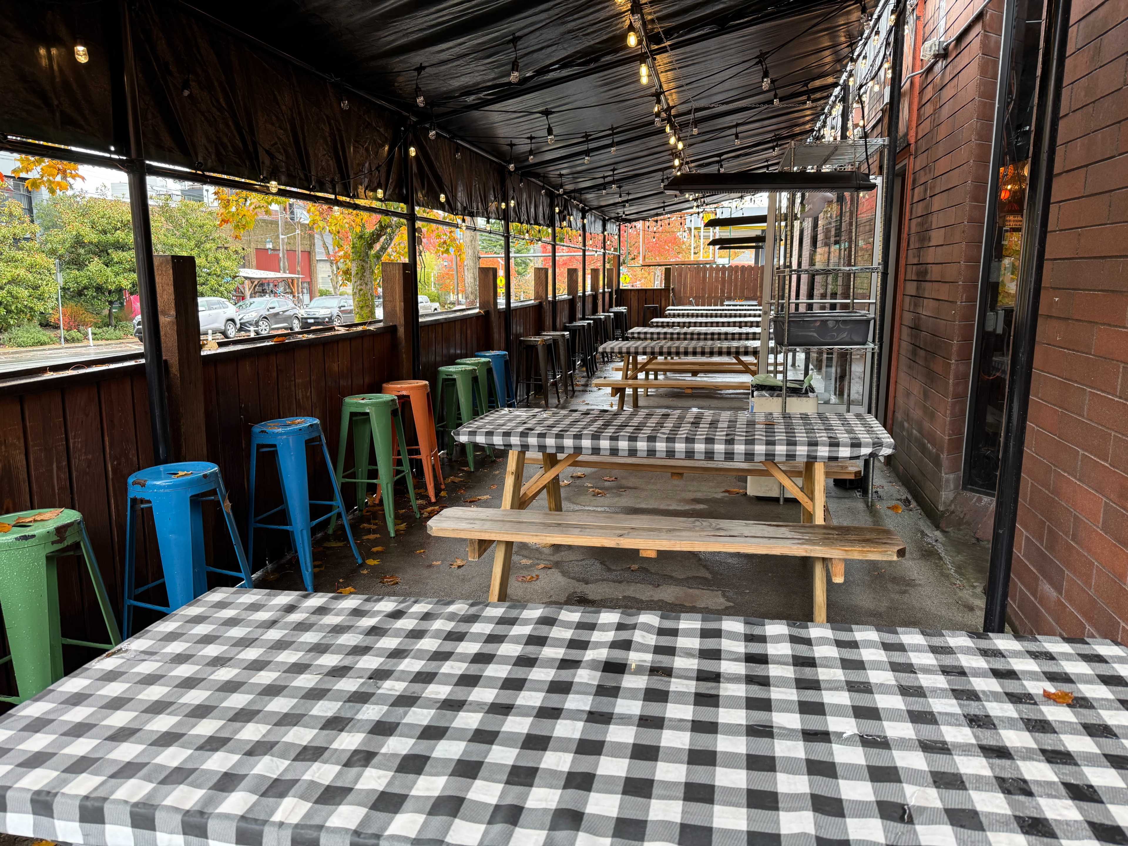 An outdoor dining area features several long tables covered with checkered tablecloths, flanked by colorful stools beneath a canopy.
