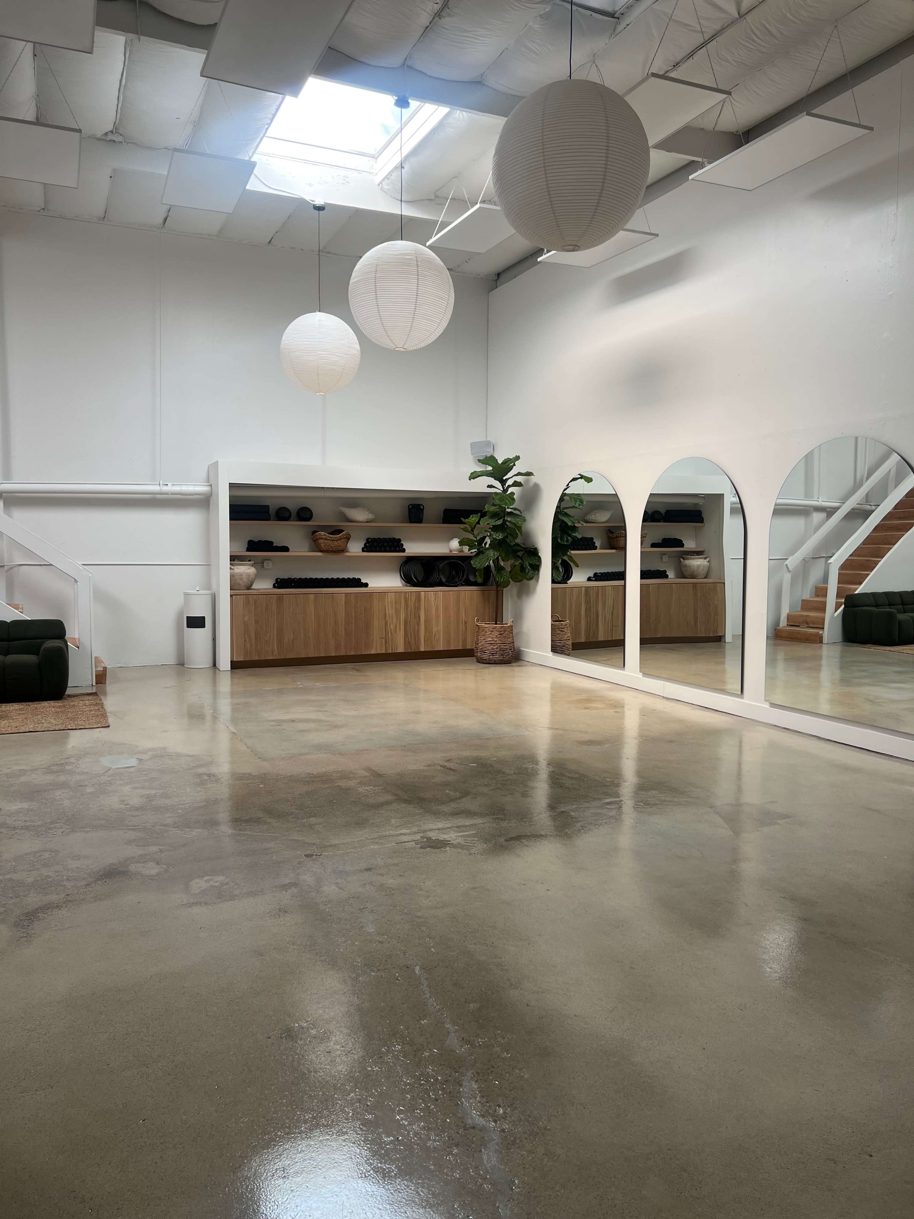 A spacious, modern studio with polished concrete floors, large mirrors, and minimalist decor featuring pendant lights and a shelf displaying rolled mats.