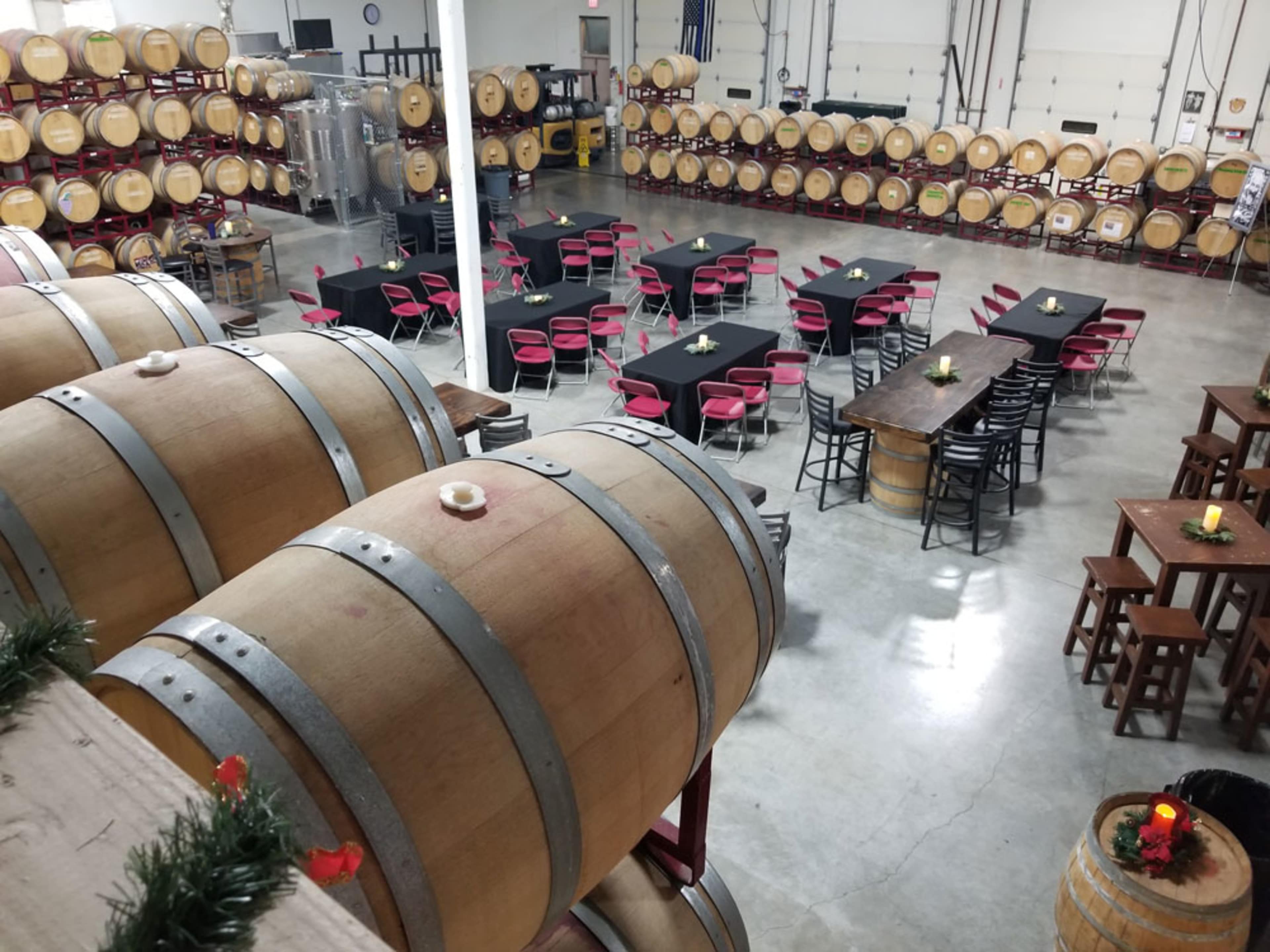 A winery interior with large wooden barrels in the foreground and tables set for dining in the background, surrounded by additional barrels lining the walls.