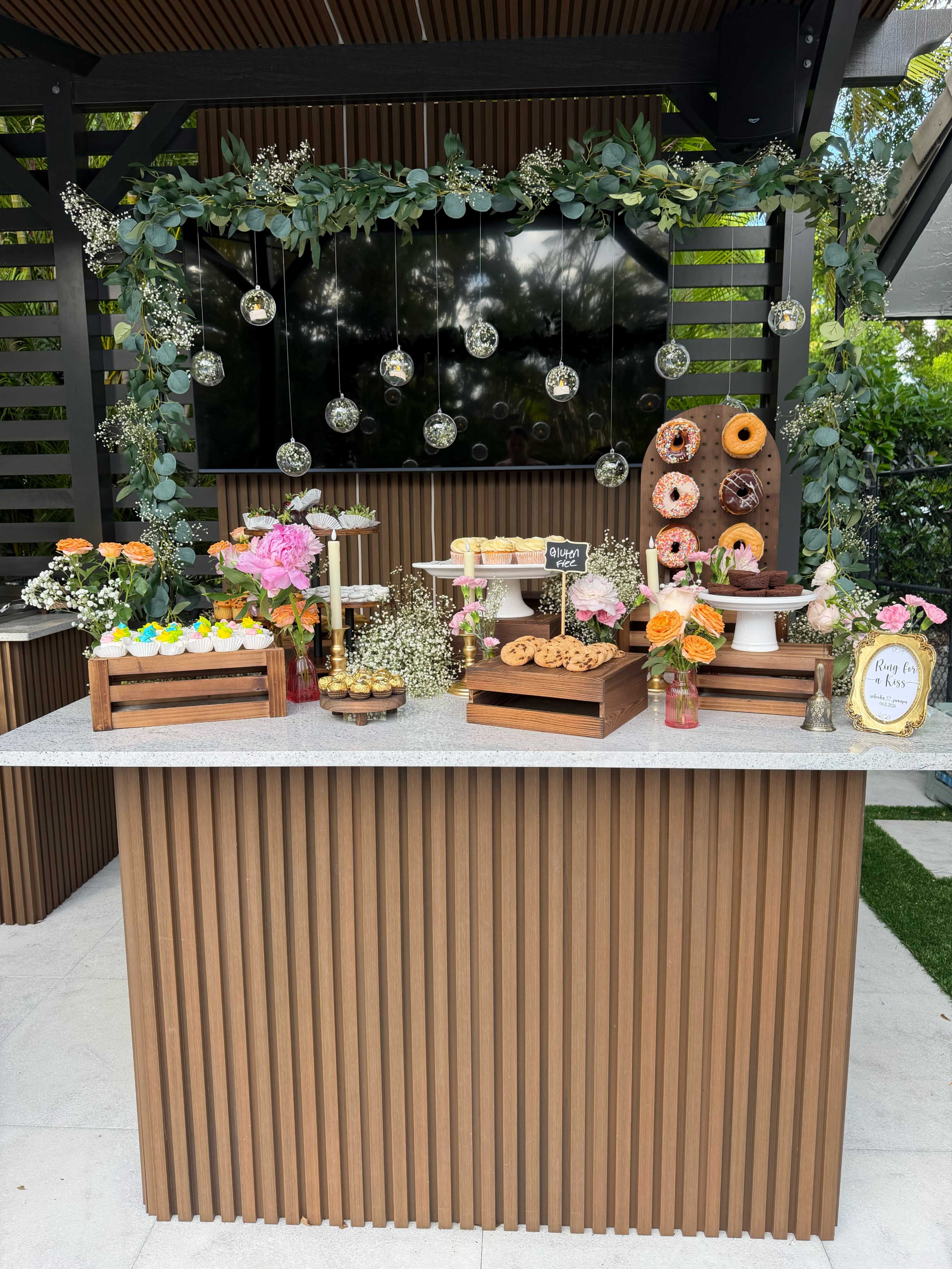 A dessert table is set up outdoors, featuring an assortment of colorful cupcakes, cookies, and donuts, surrounded by floral arrangements and decorative greenery.