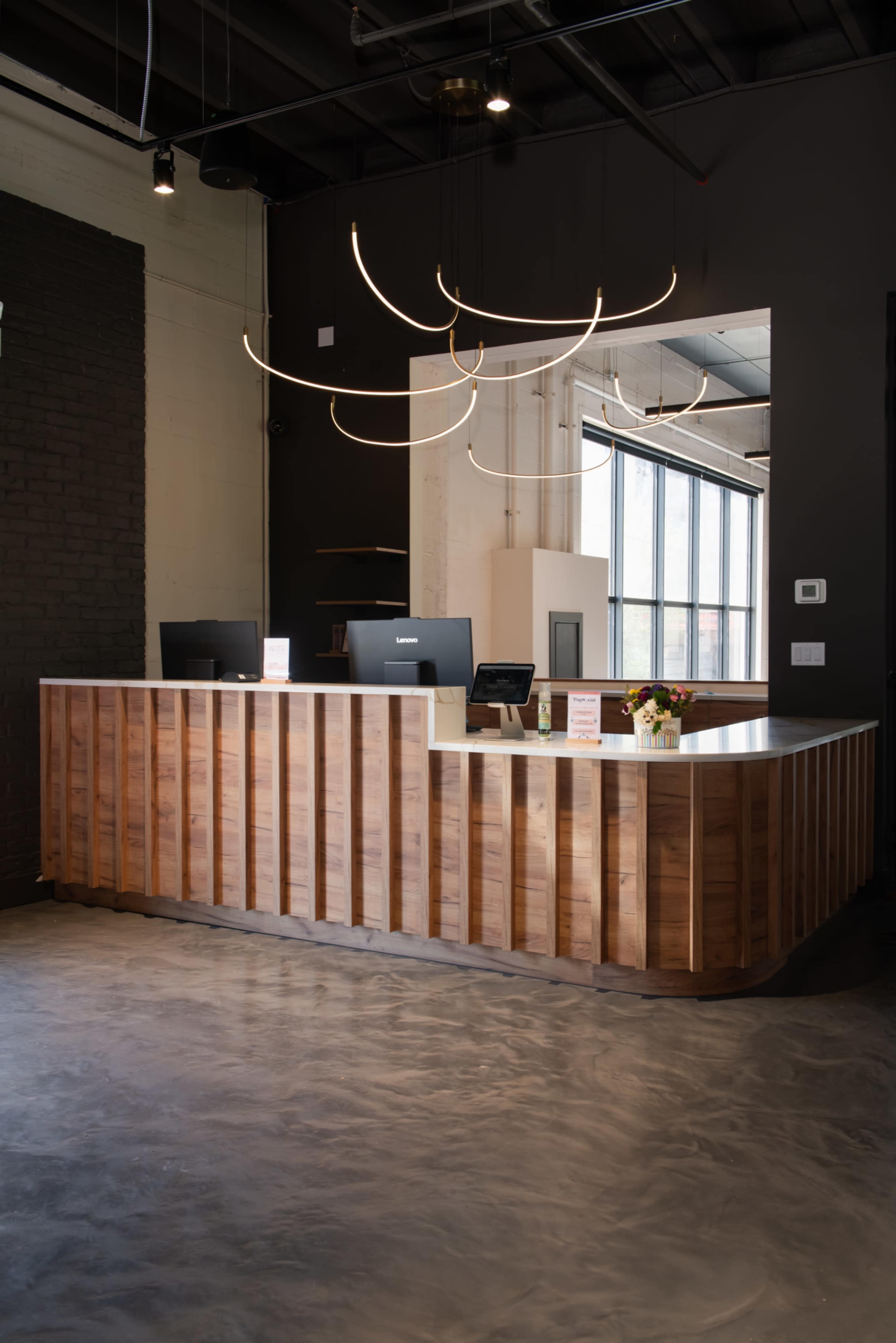 The image shows a modern reception desk made of wood, positioned in a well-lit space with large windows and a minimalist design featuring stylish light fixtures.