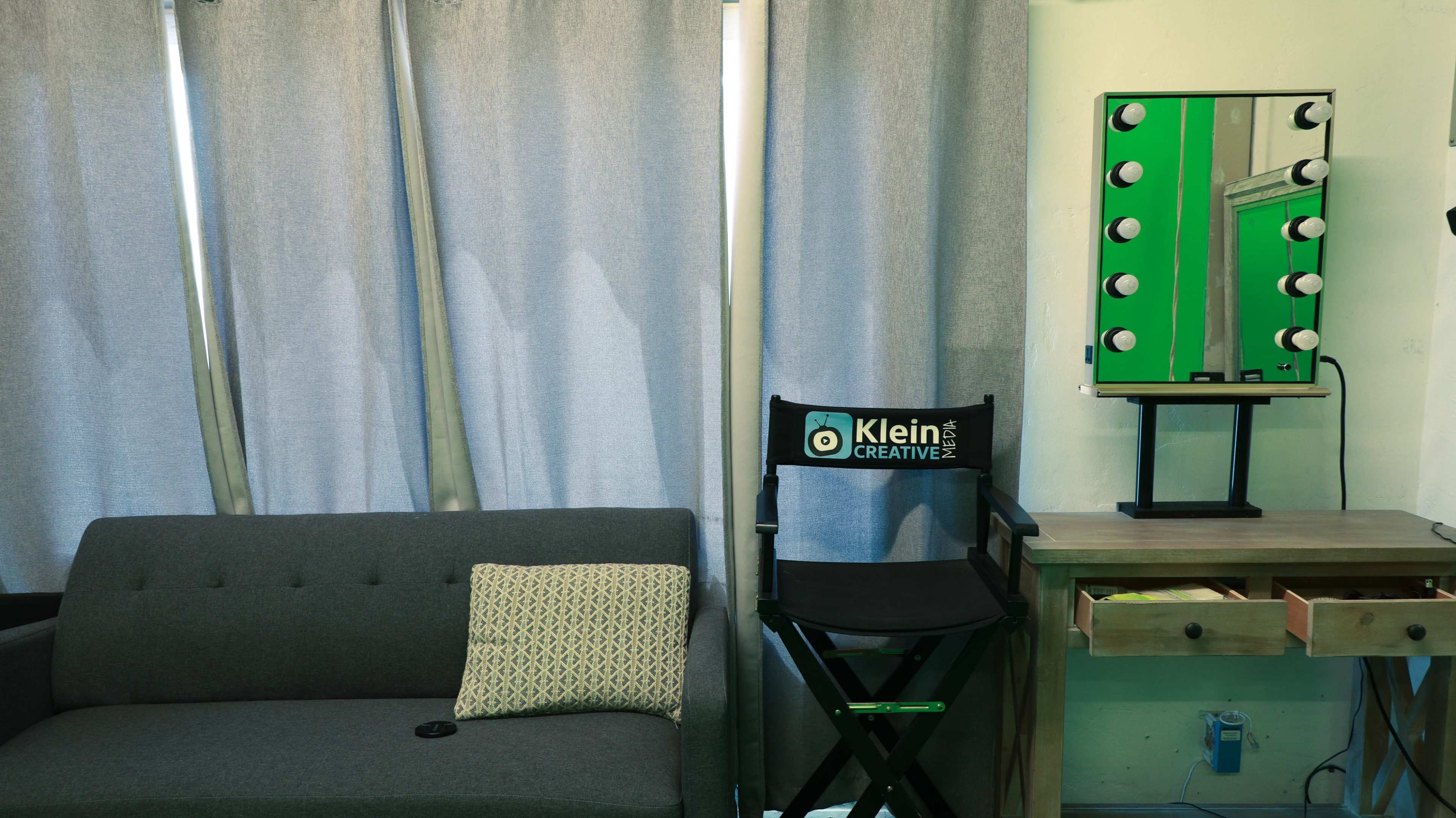 The scene depicts a minimalist indoor space featuring a gray sofa with a decorative pillow, a director's chair branded with "Klein Creative," and a lit vanity mirror against a green wall.