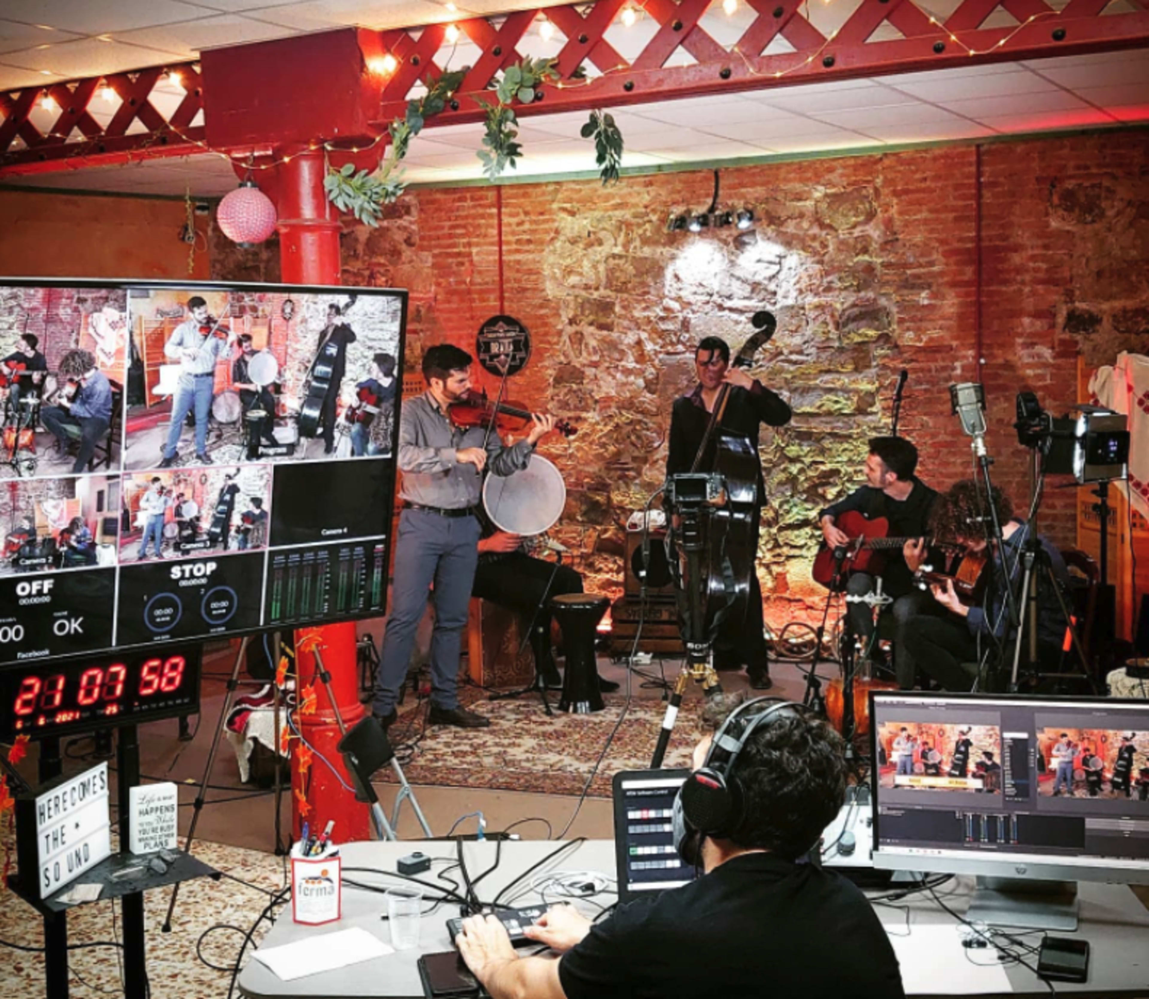 A group of musicians performs in a rustic room equipped with cameras and monitors for a live-streamed event.