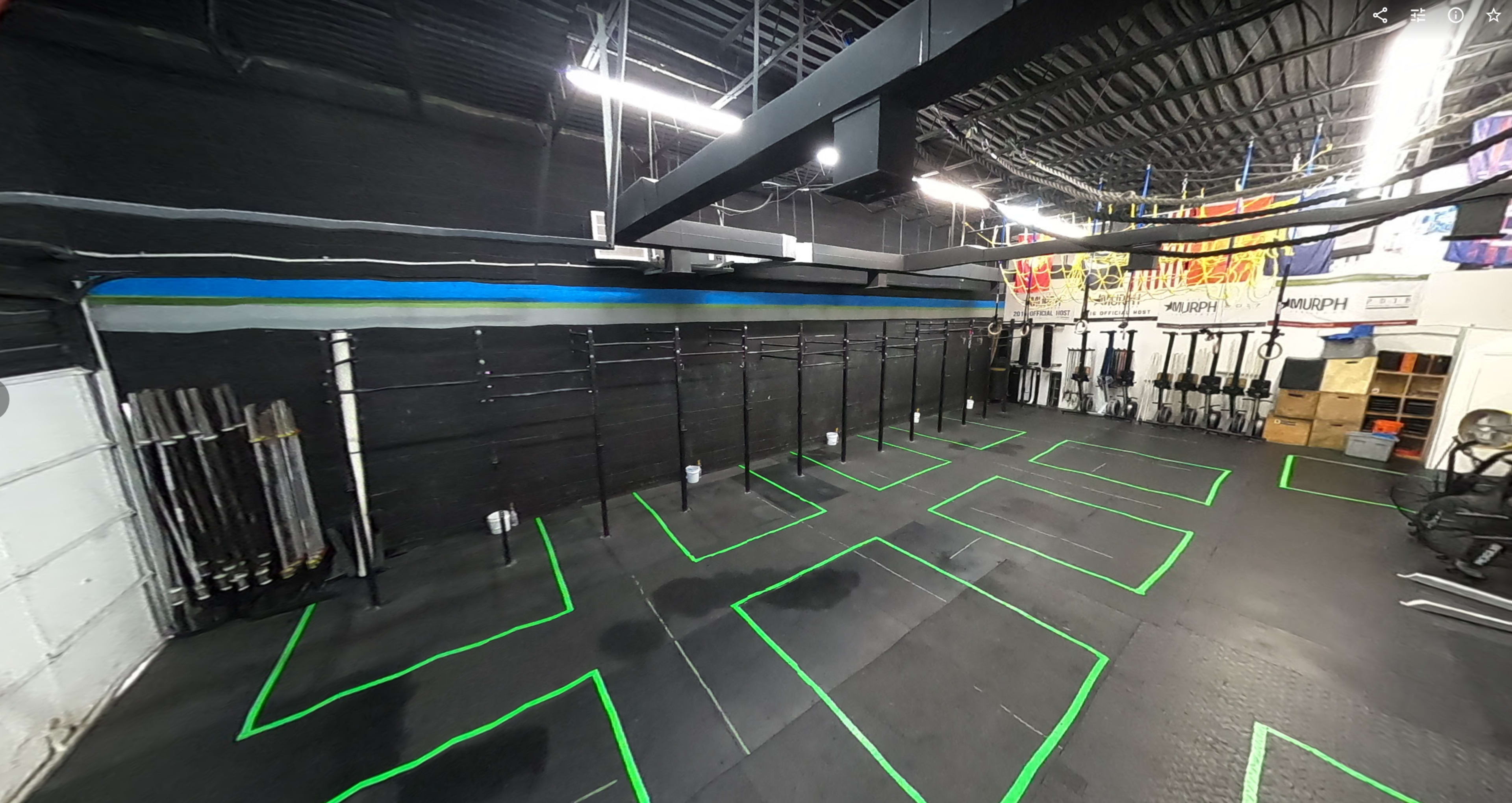 A gym interior with black walls, workout equipment arranged along the back, and green floor markings for exercise stations.