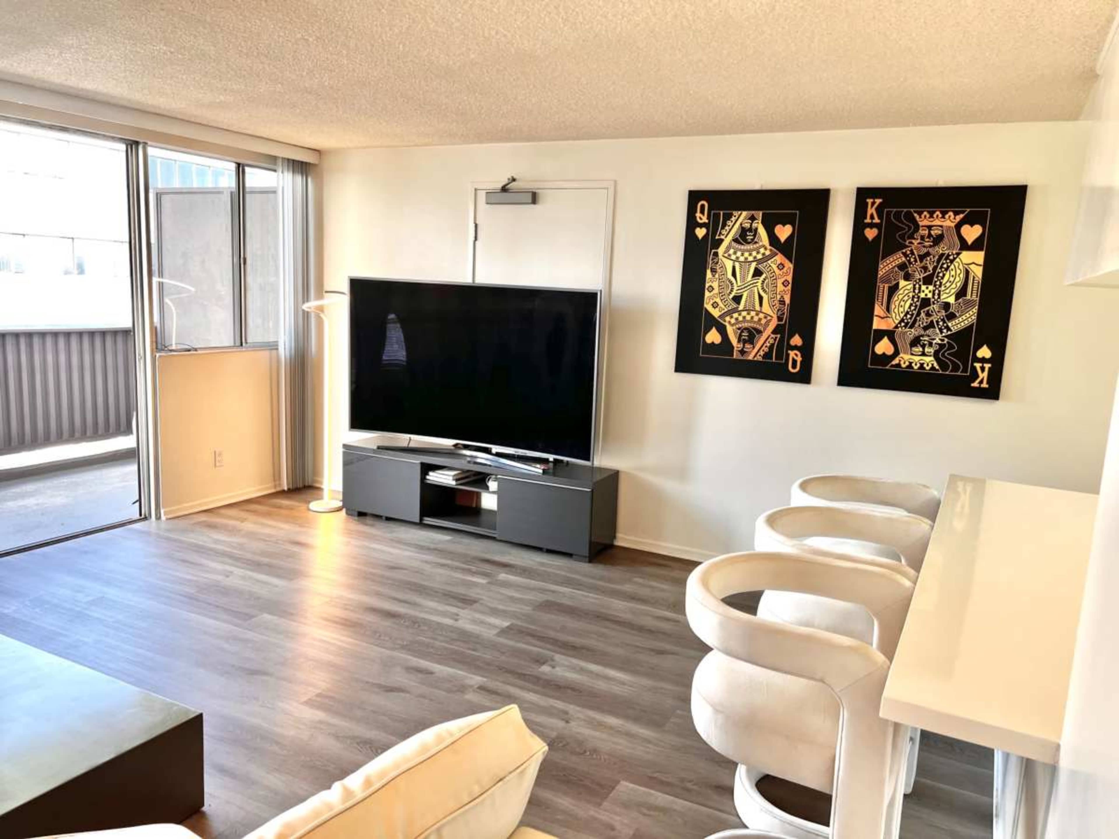 The image shows a modern living room with a large television on a low cabinet, two framed artwork pieces on the wall, and a bar area with white stools.