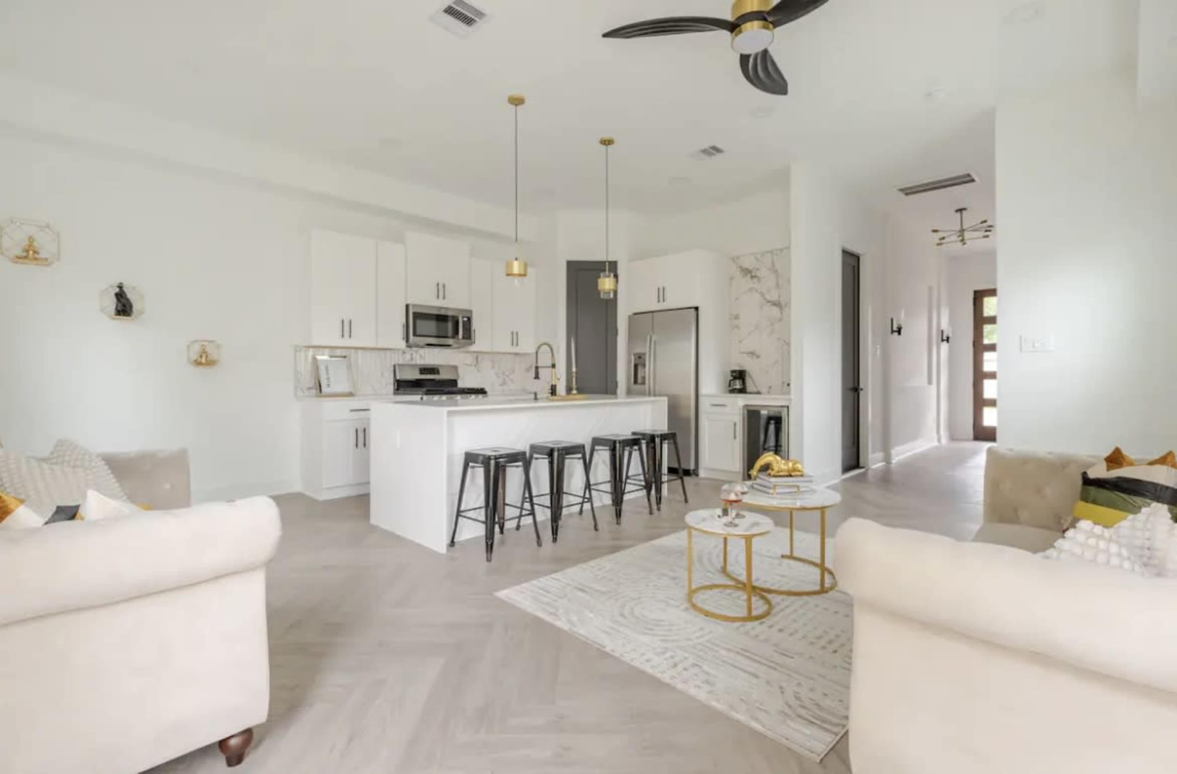 A modern kitchen and living area feature white cabinetry, stainless steel appliances, and a spacious island with bar stools.