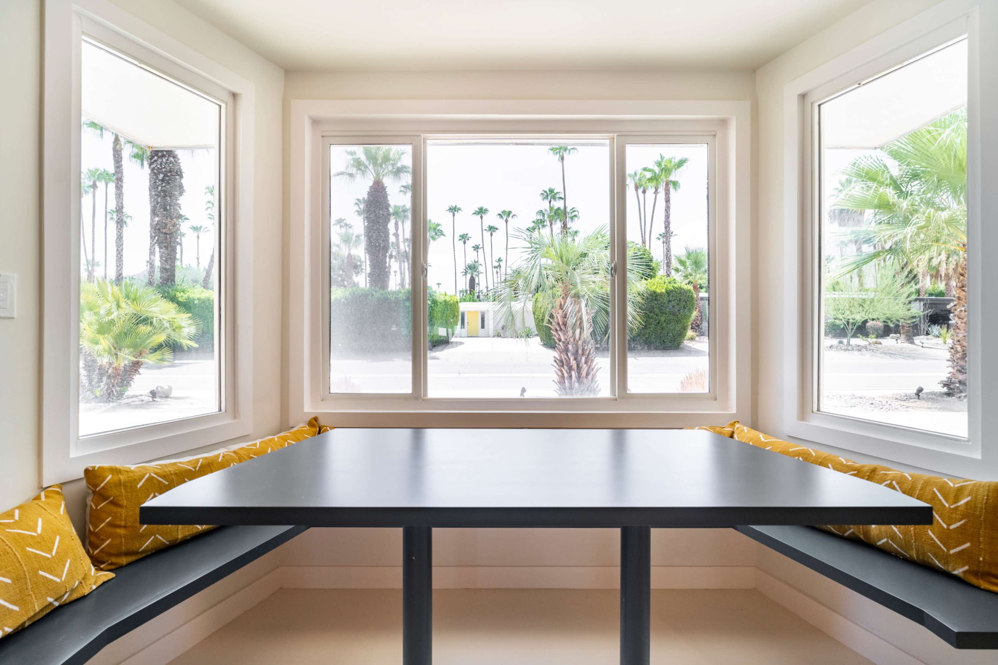 A modern corner dining area features a black table surrounded by windowed walls showcasing palm trees outside.