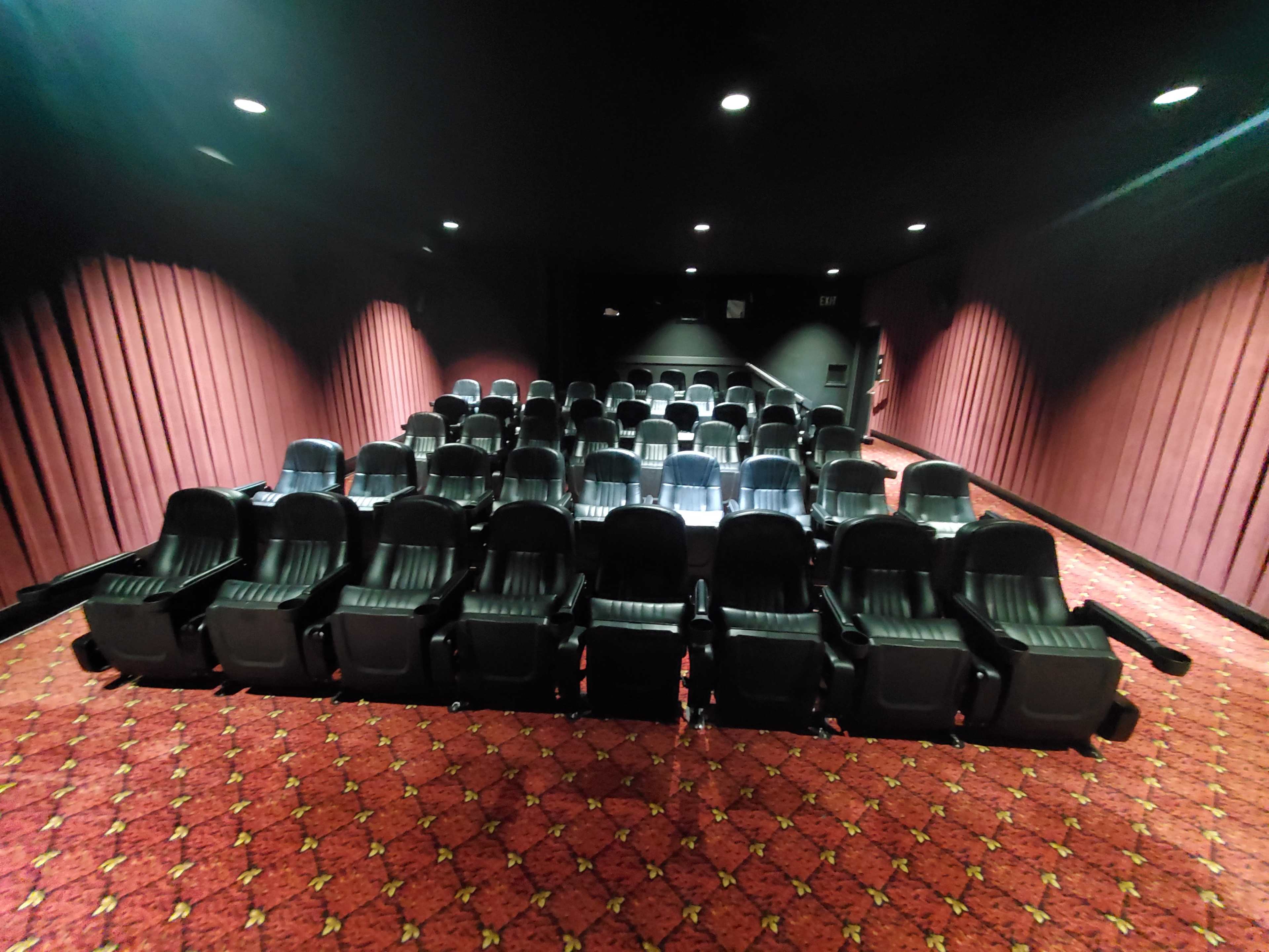 The image shows a darkened movie theater with rows of empty black reclining seats arranged on a patterned carpet.