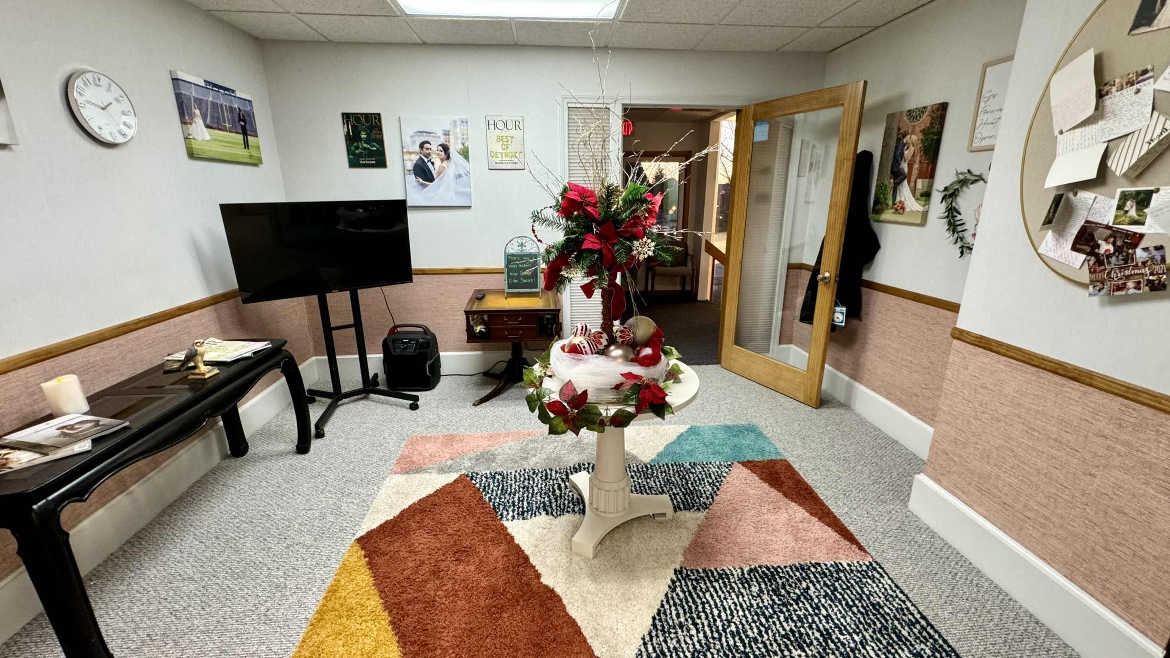 The image shows a small room with a colorful rug, a round table decorated with flowers, a television, and various wall decorations.