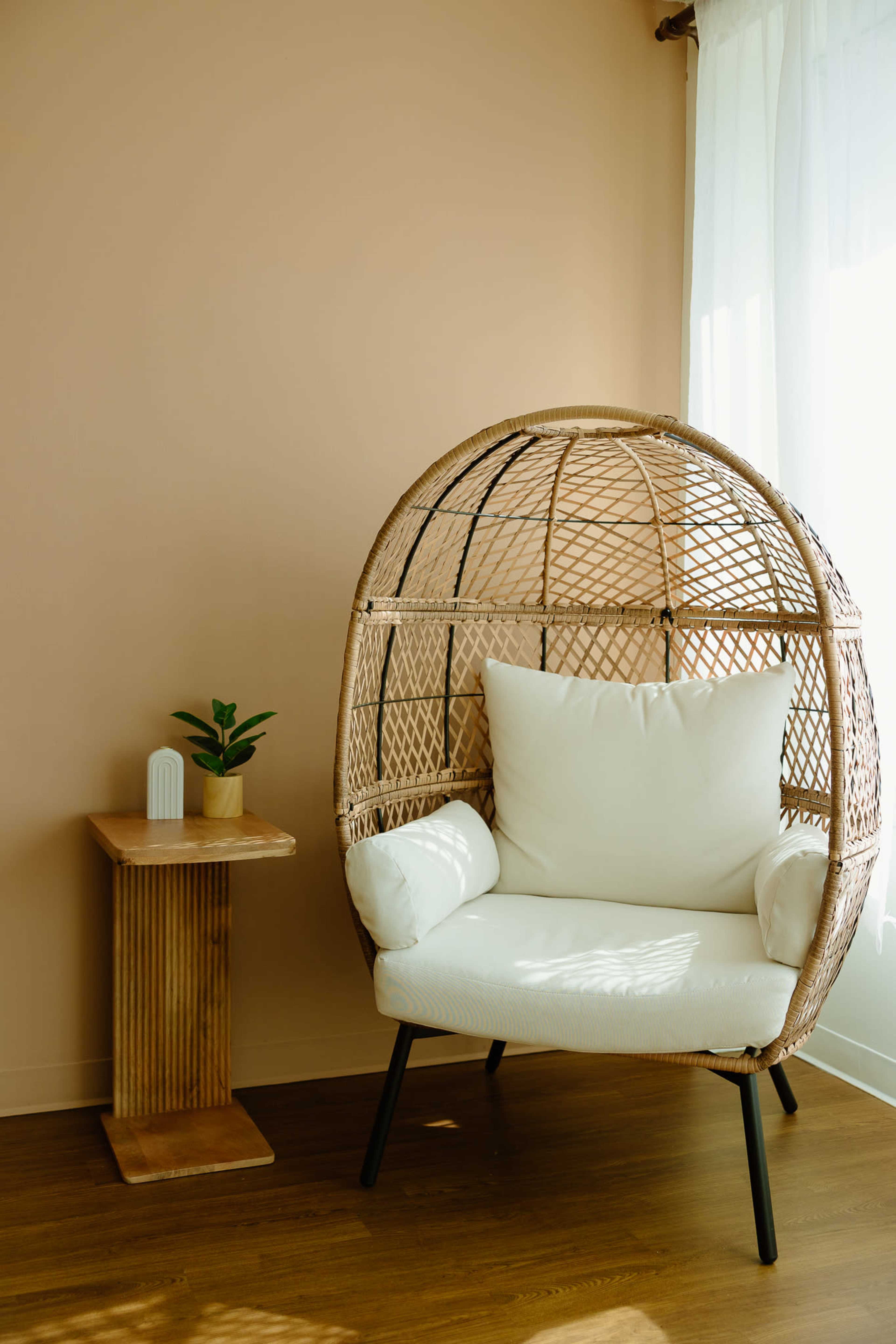 A woven rattan chair with a white cushion sits next to a wooden side table and a potted plant against a light beige wall.