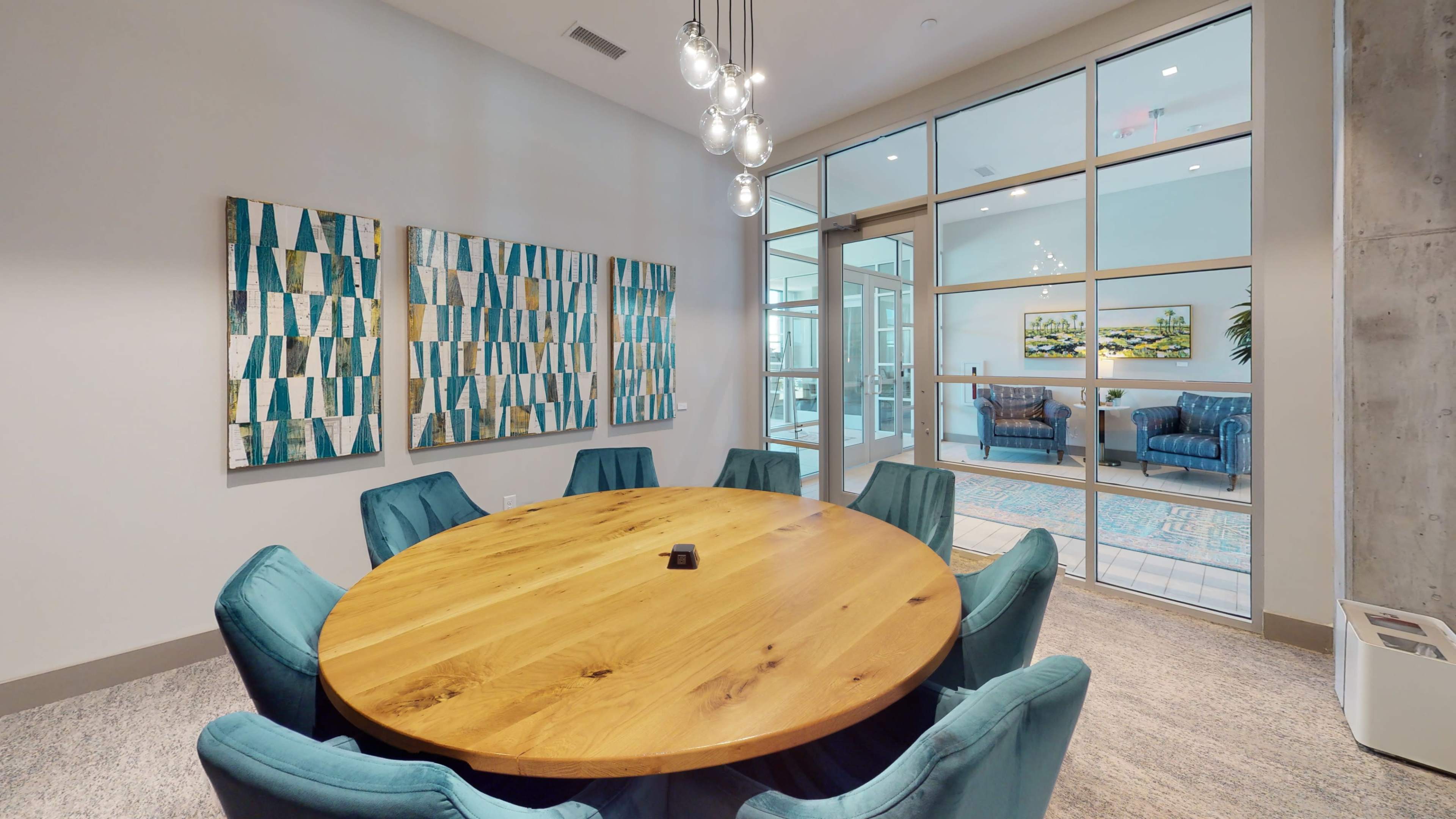 A modern meeting room featuring a large round wooden table surrounded by teal chairs, with abstract artwork on the walls and glass doors leading to a lounge area.