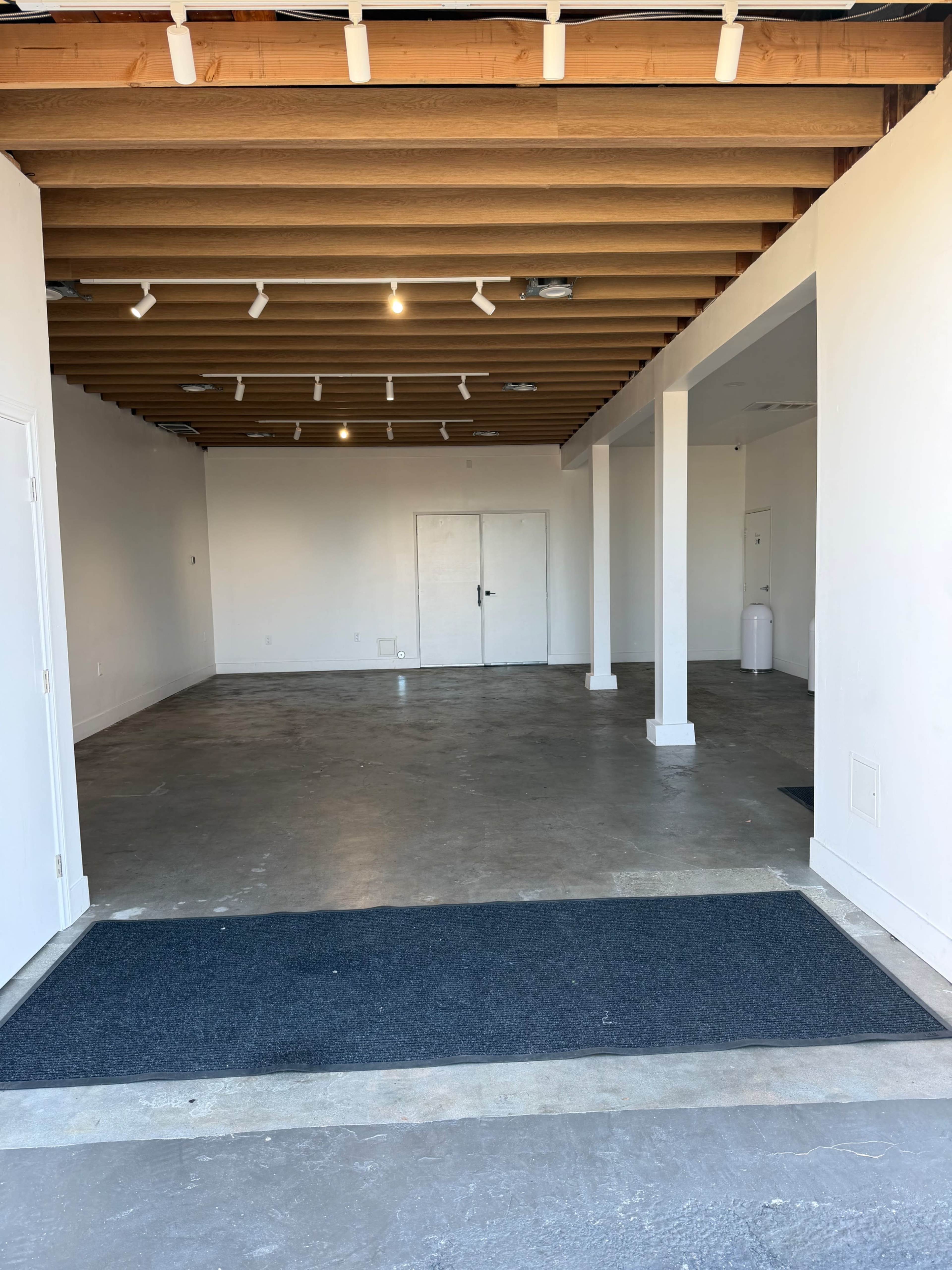 The image shows a spacious, empty interior with exposed wooden beams, concrete flooring, and two sets of double doors at the back.