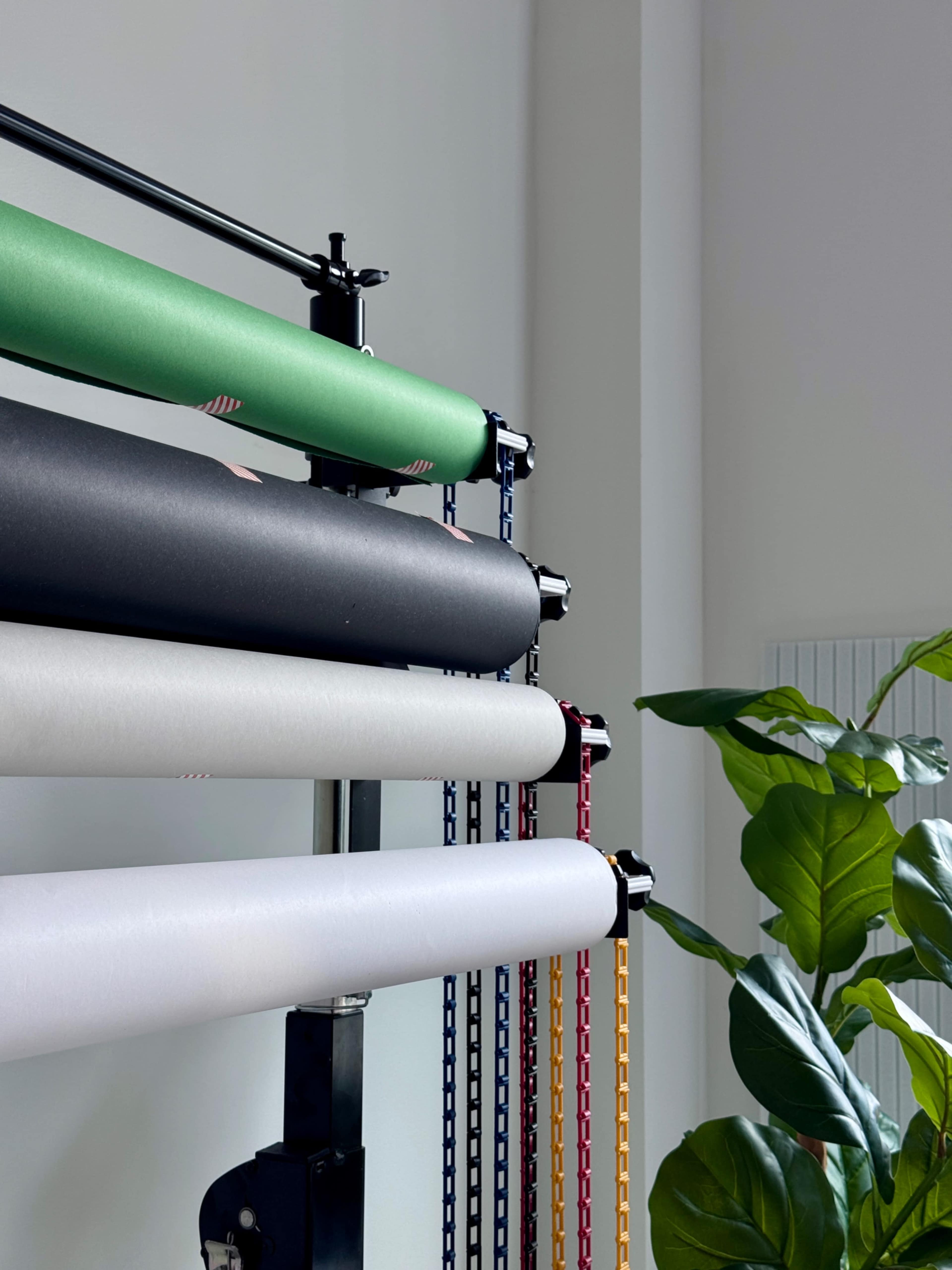 The image features a vertical display stand holding multiple rolls of colored paper alongside a potted plant in a bright room.