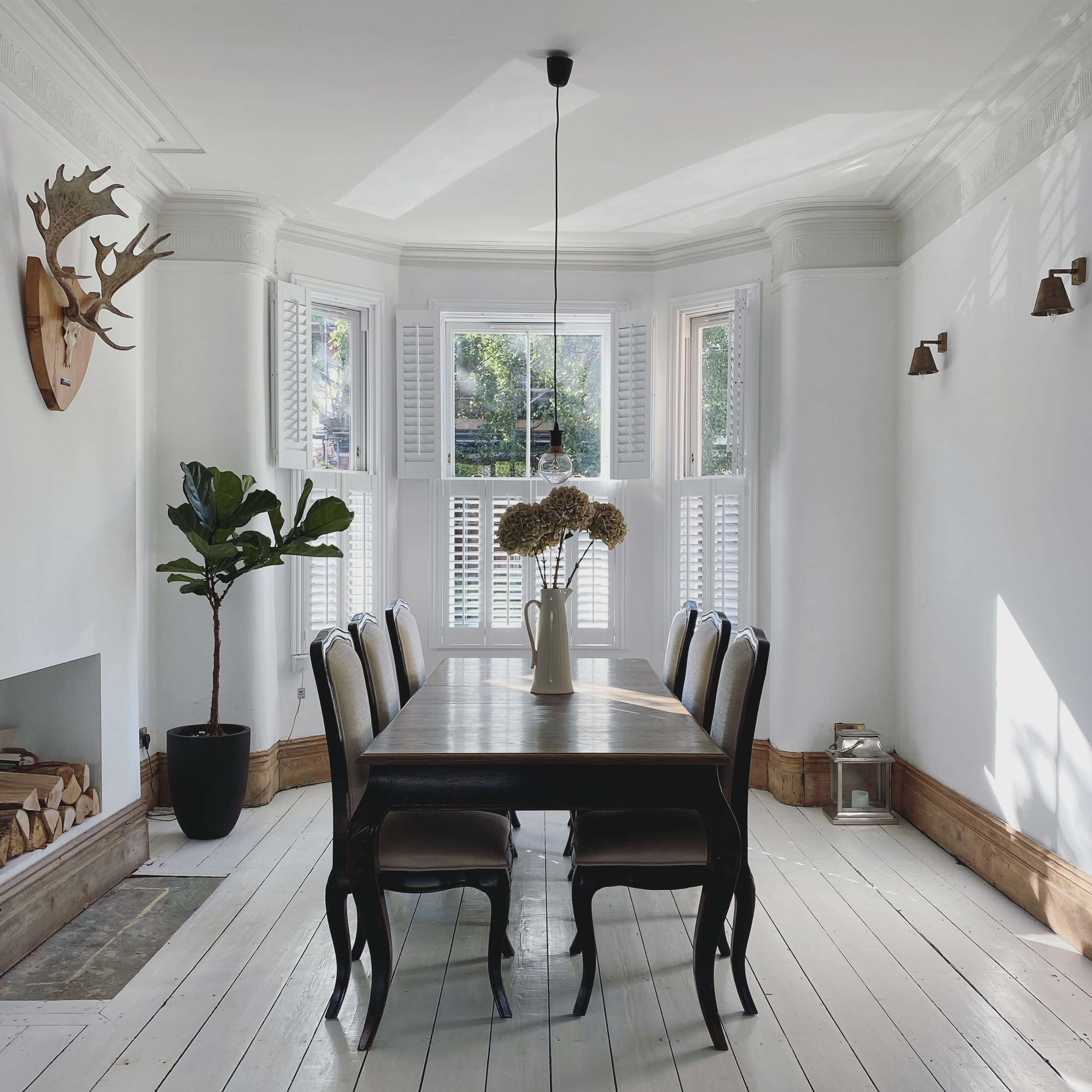 A dining room features a long wooden table surrounded by six chairs, with a potted plant in one corner and large windows allowing natural light.