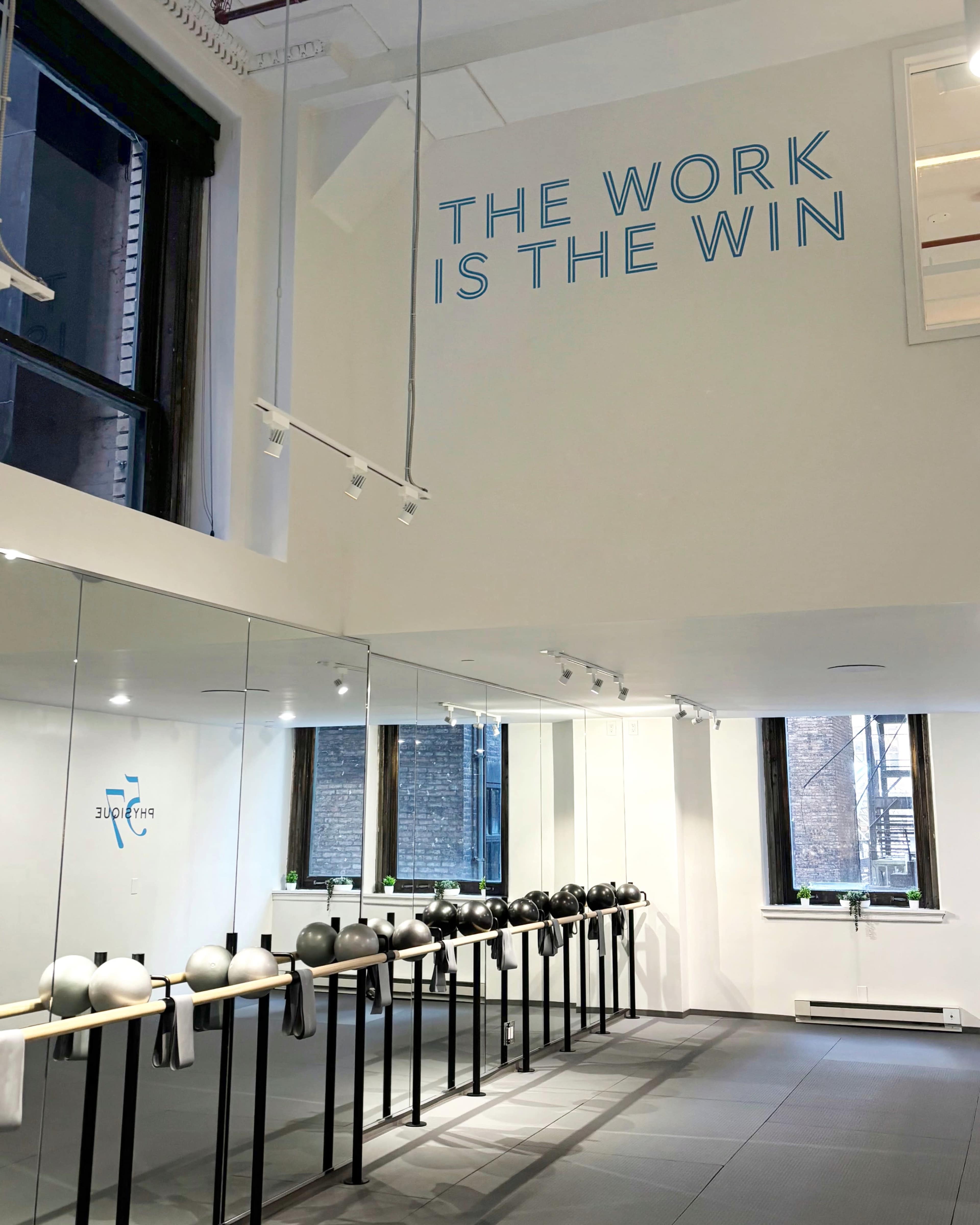 The image shows an interior fitness studio with a row of black exercise equipment along a mirrored wall, and the phrase "THE WORK IS THE WIN" displayed on the white wall above.
