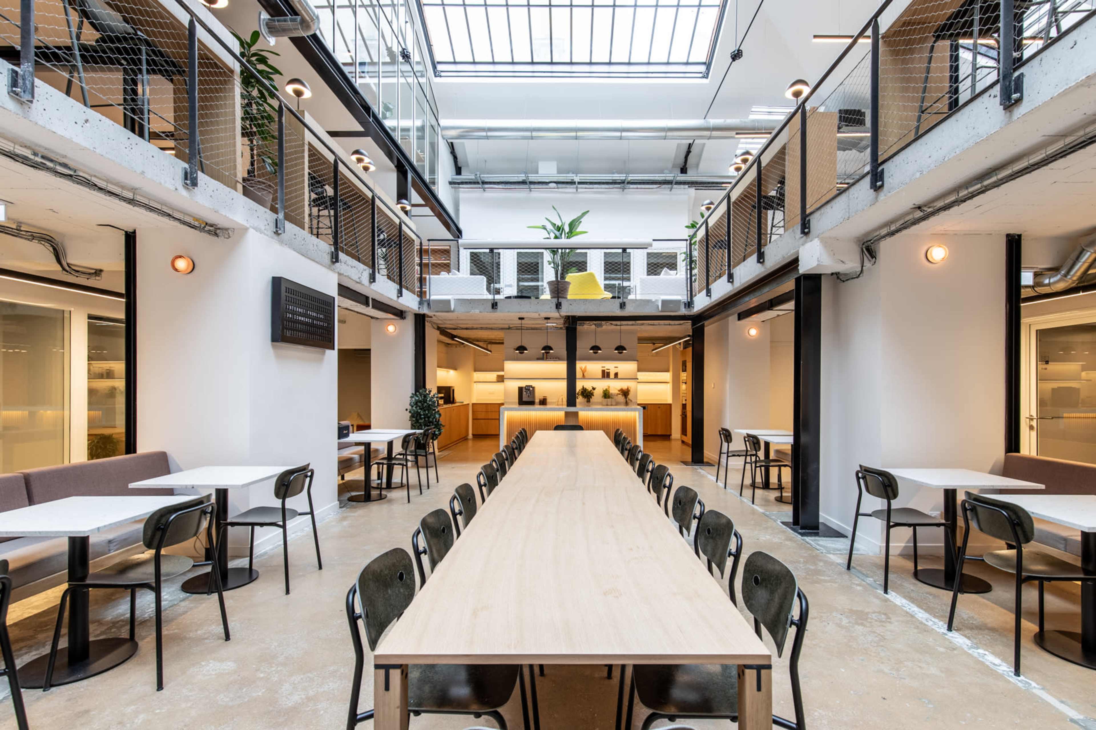 The image shows a spacious, modern cafeteria with a long central table, surrounded by chairs, open to a bright area with plants and overhead skylights.