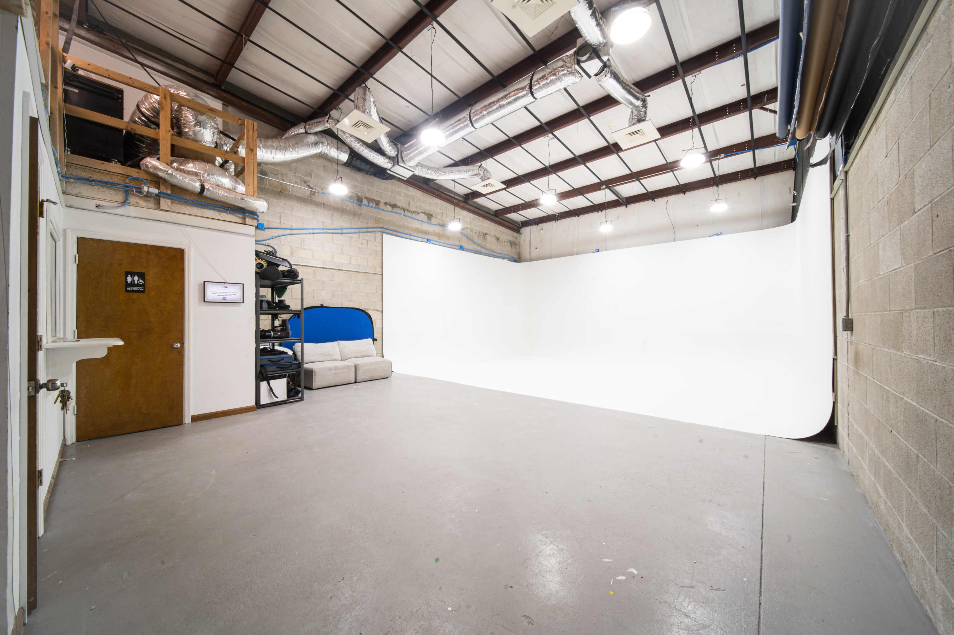 The image shows an empty photography studio with a white cyclorama wall, a gray floor, and a few basic furnishings including a sofa and storage shelves.
