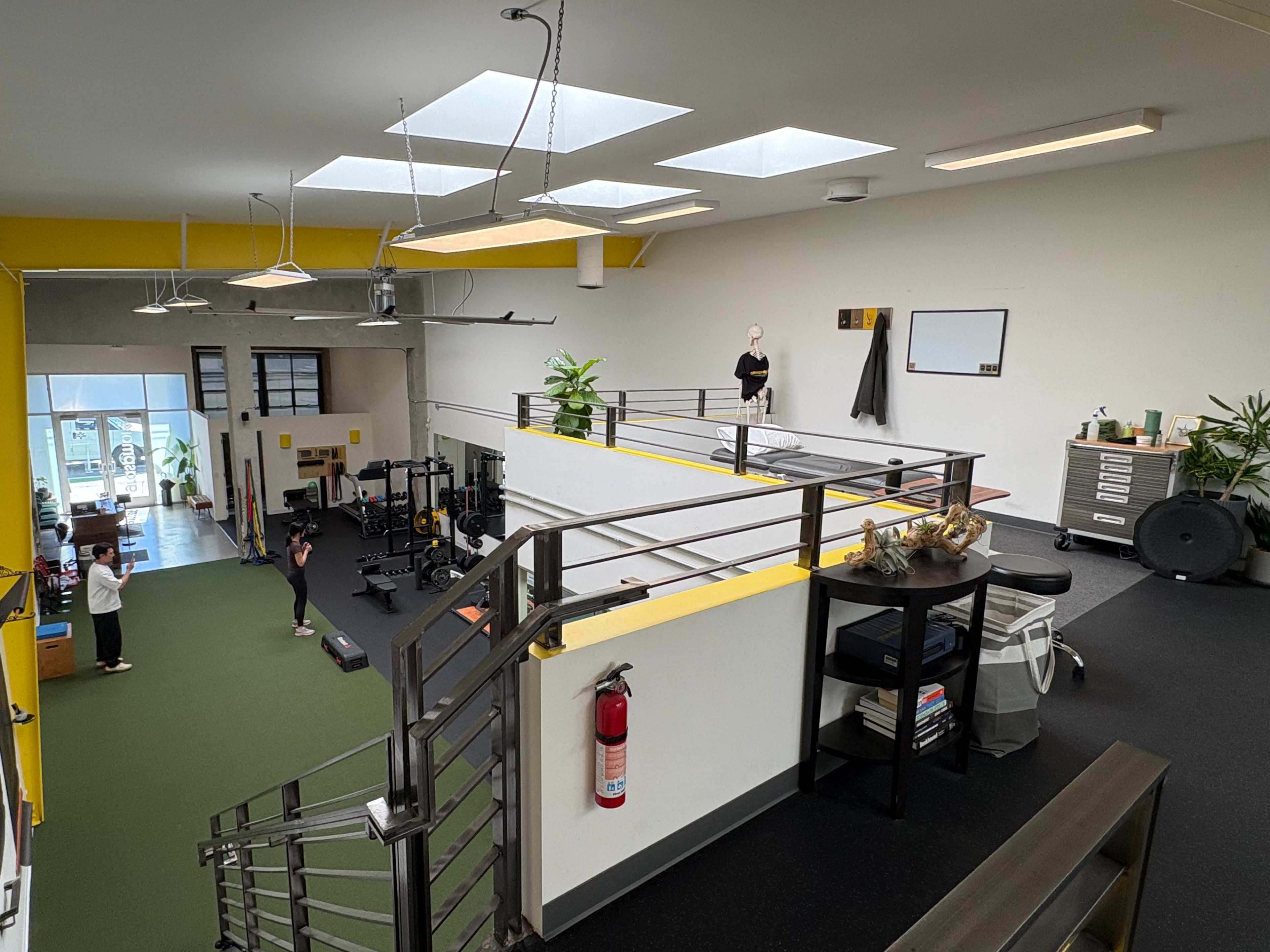 The image shows a spacious gym with various workout equipment, natural light from skylights, and a balcony area overlooking the workout space below.