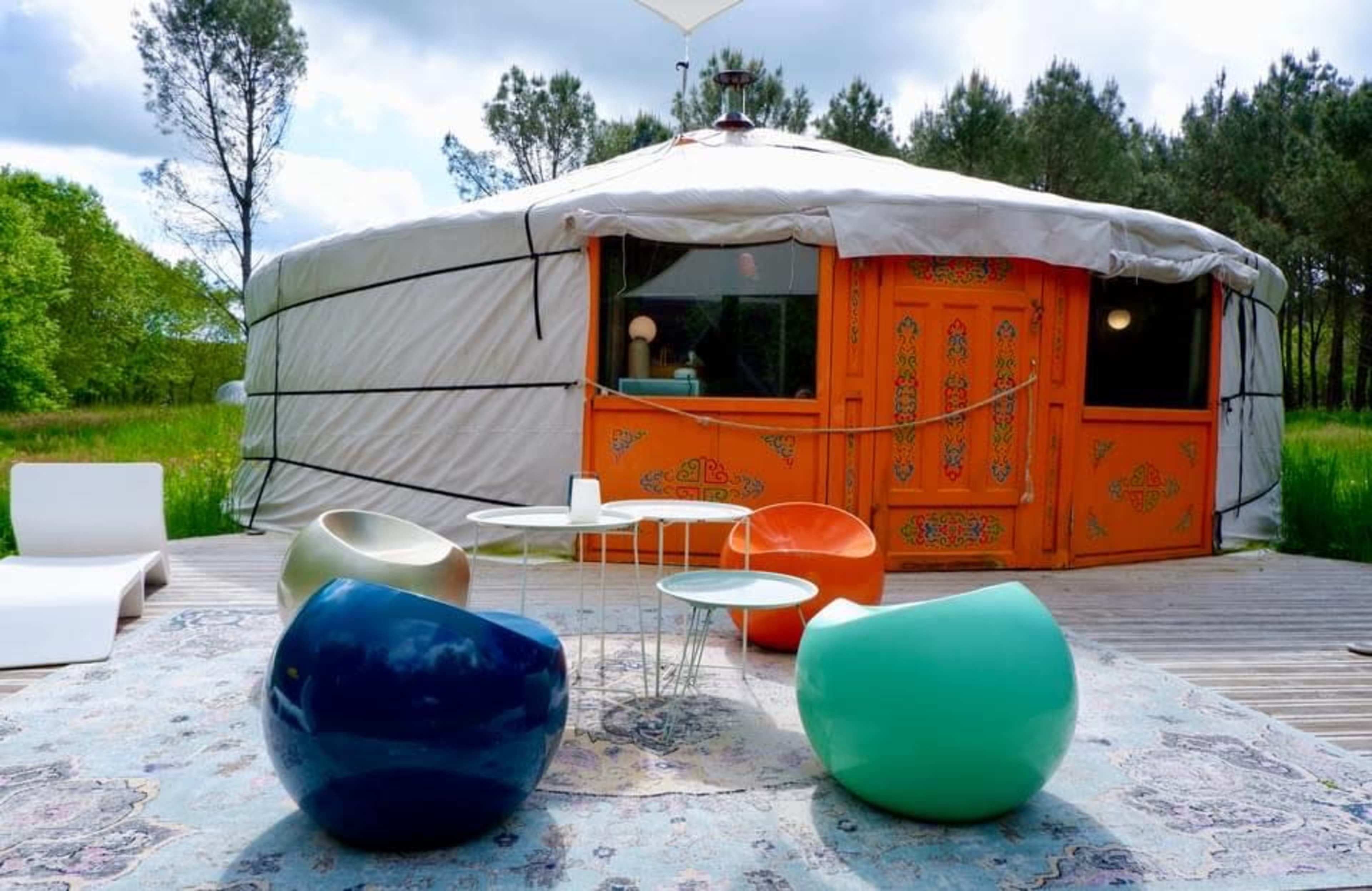 A colorful seating area with modern chairs and a table is set on a wooden deck in front of a traditional yurt surrounded by trees.