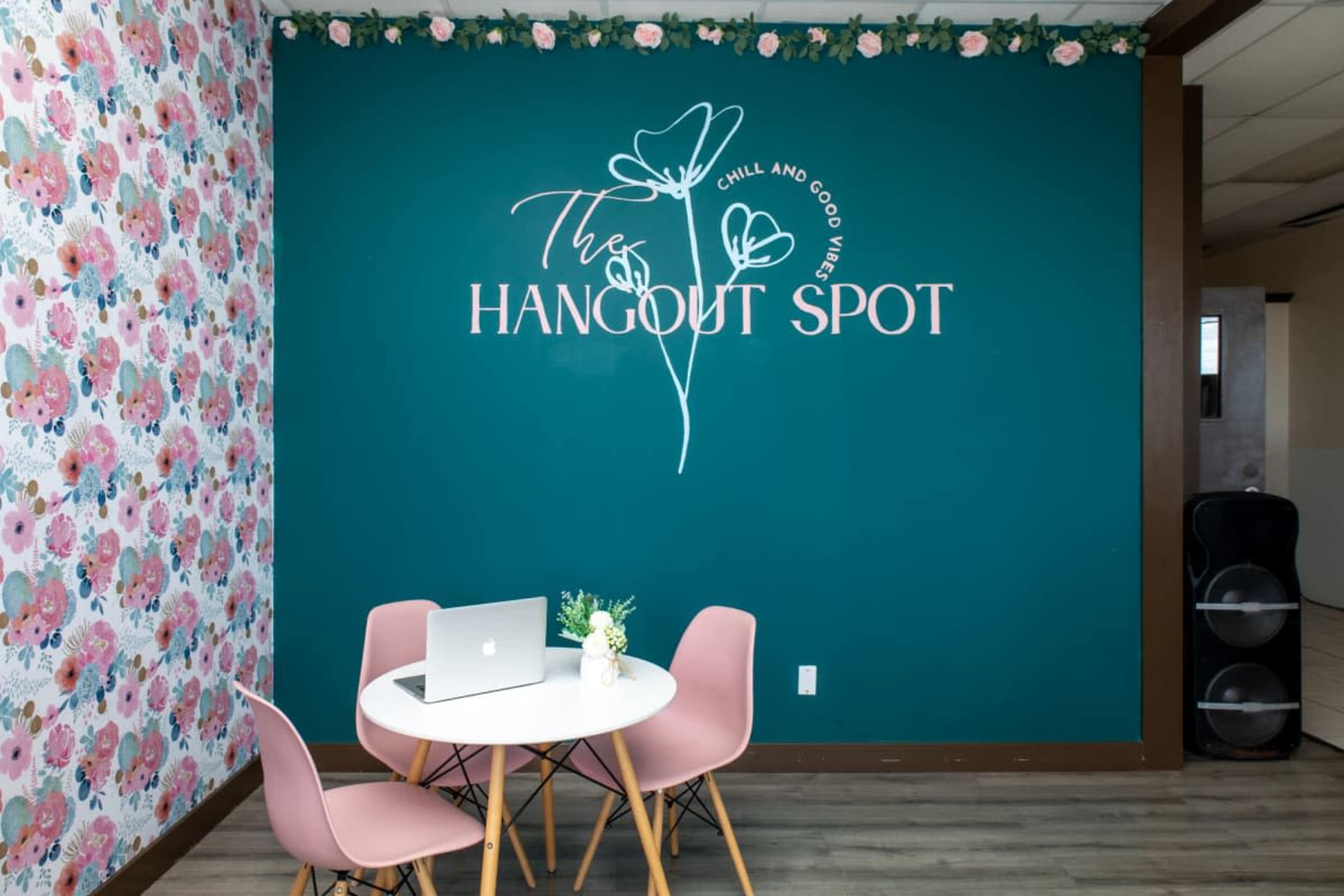 The image shows a small indoor space with a round white table and pink chairs, featuring a teal wall with a decorative sign that reads "The Hangout Spot" and floral wallpaper on one side.