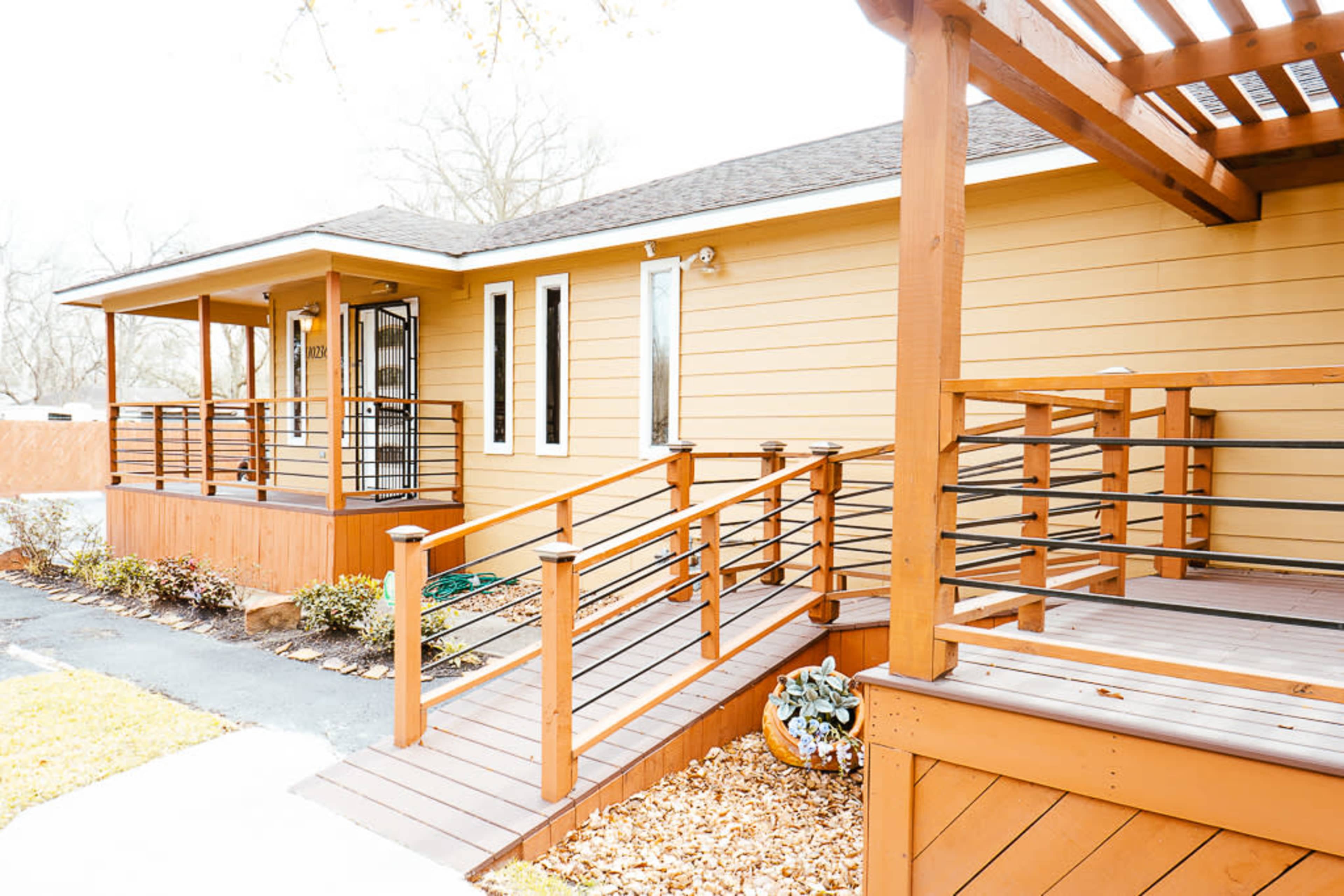 A single-story house with a wooden deck and railing is shown, featuring a pathway leading to the entrance.