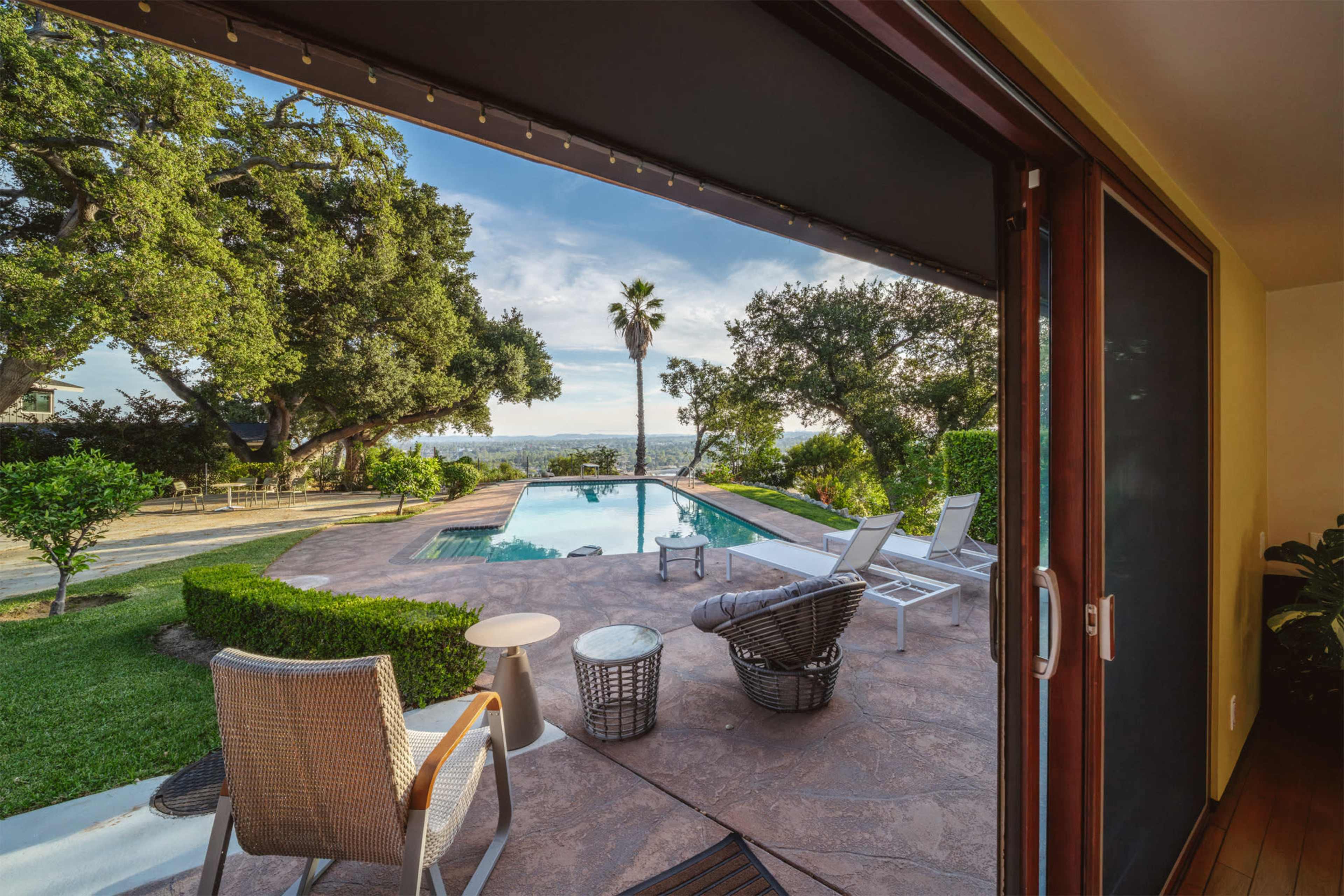 The image shows a backyard view of a pool surrounded by lounge chairs and lush greenery under a blue sky.