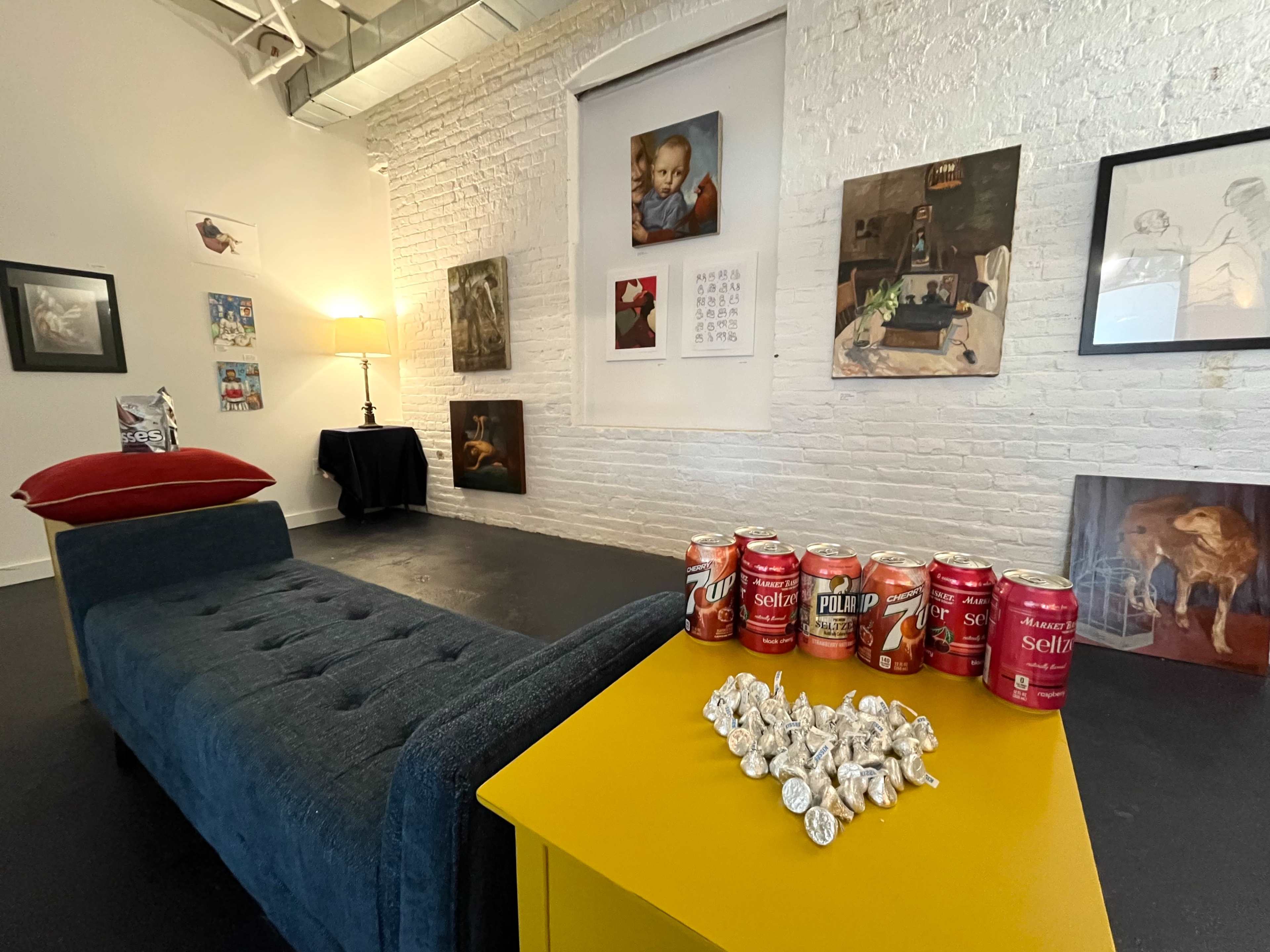 A modern art gallery features a blue chaise lounge, a yellow side table with soda cans and wrapped candies, and various framed artworks on the white brick walls.
