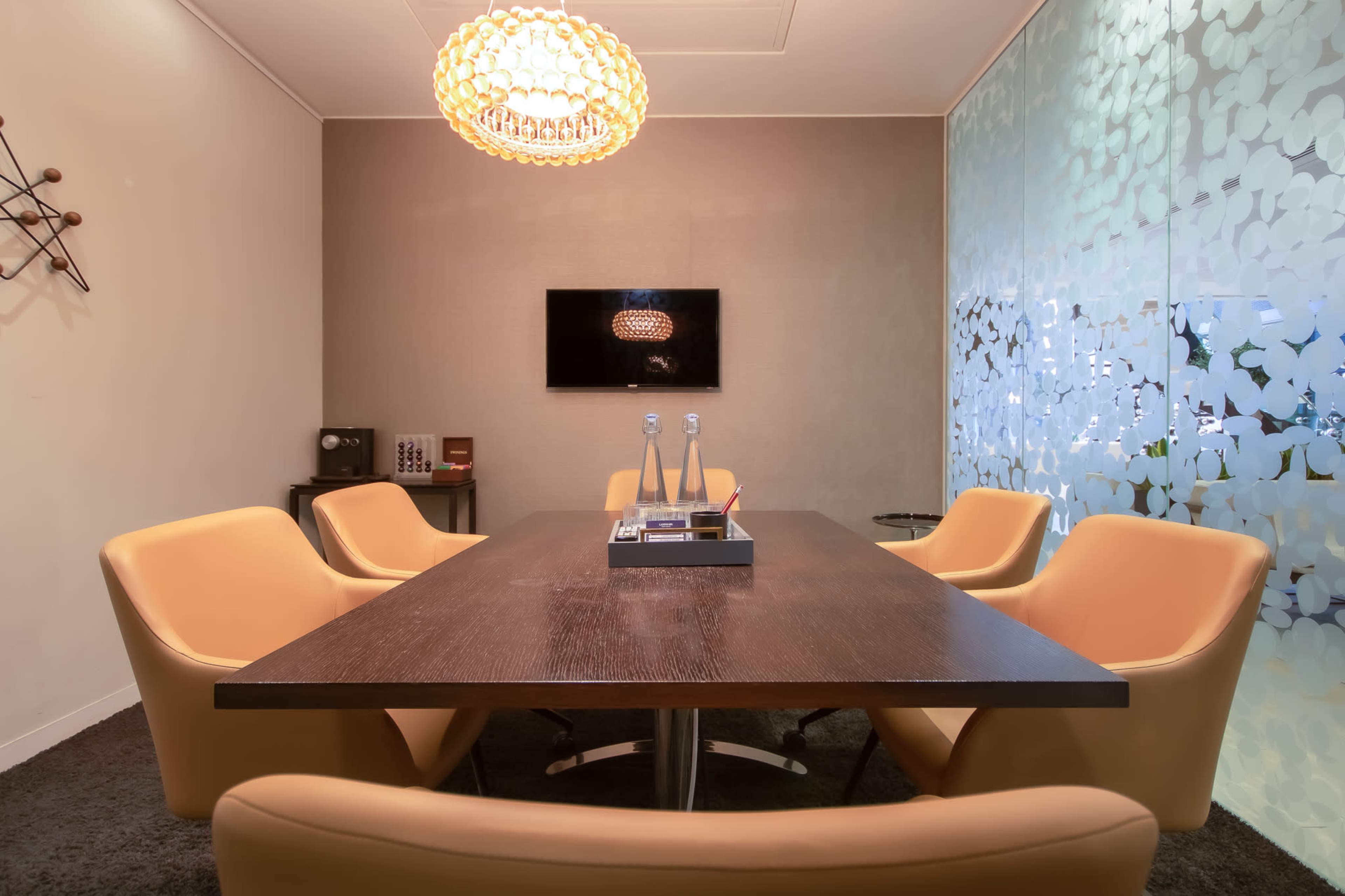 A well-furnished conference room features a large wooden table surrounded by orange chairs, a modern chandelier overhead, and a wall-mounted television.