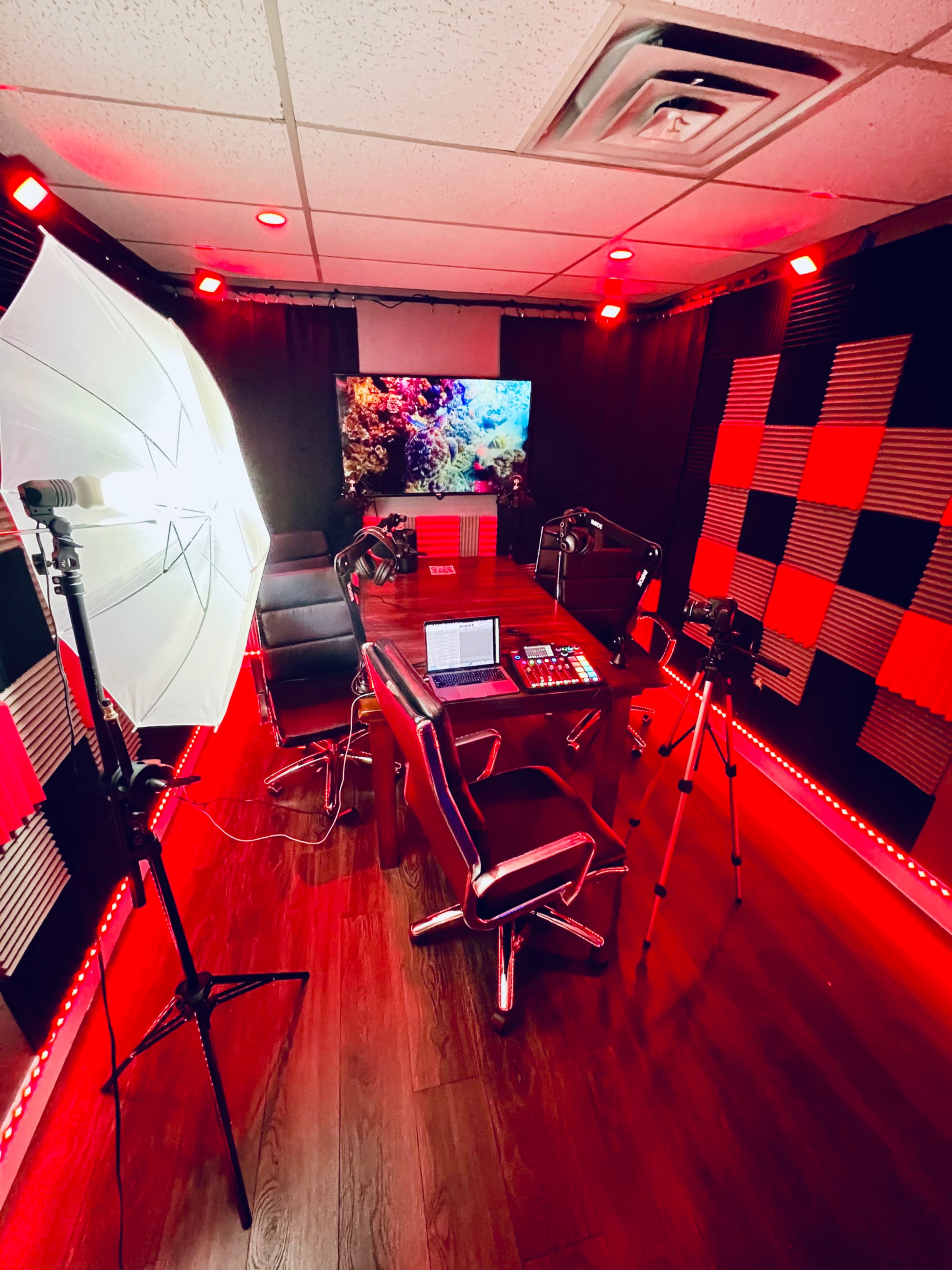 A recording studio setup with a table, chairs, a computer, and lights, surrounded by checkered wall patterns and a large screen displaying colorful visuals.