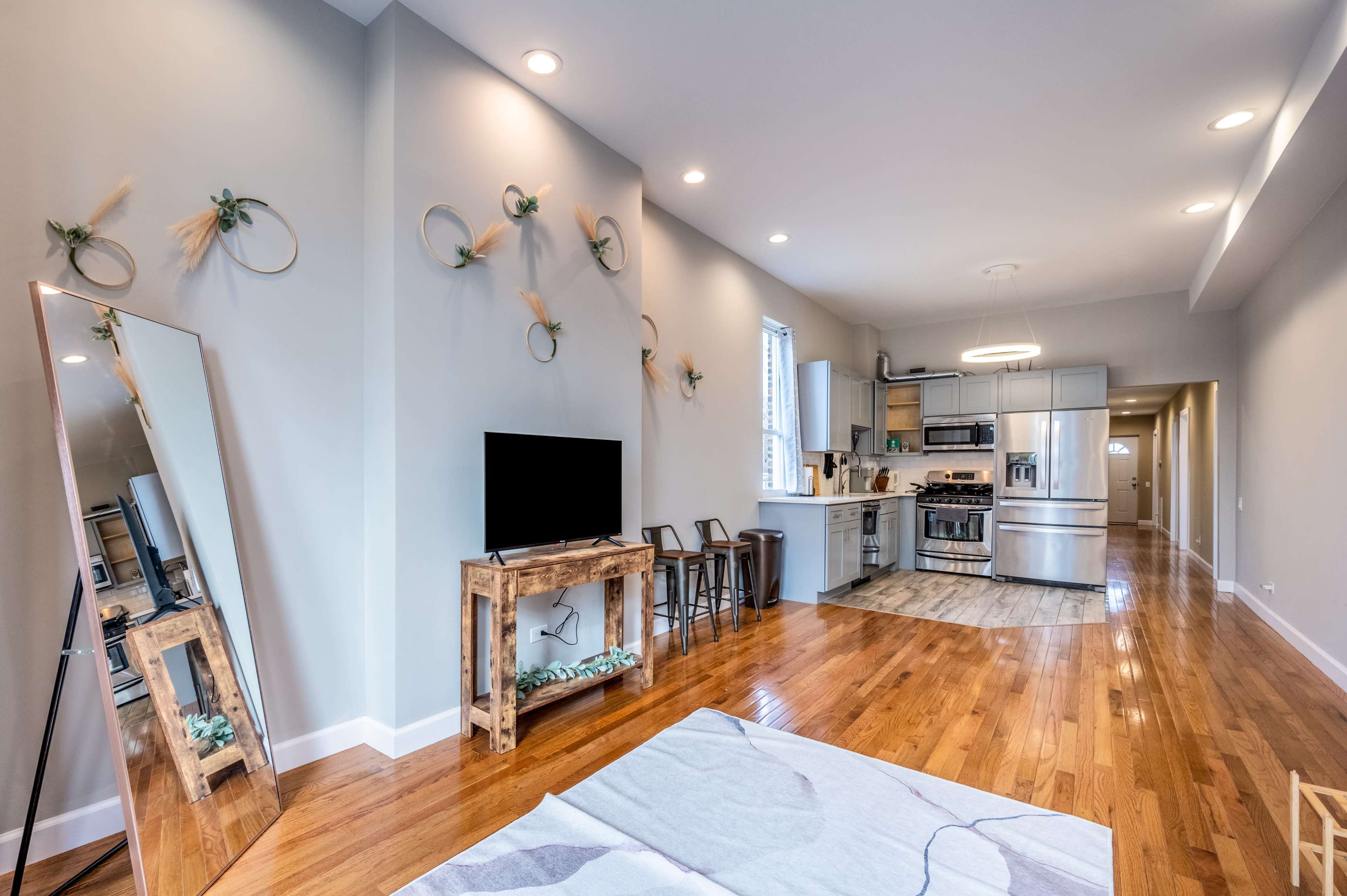A modern living space featuring a kitchen area, wooden flooring, a television on a wooden stand, and decorative wall accents.