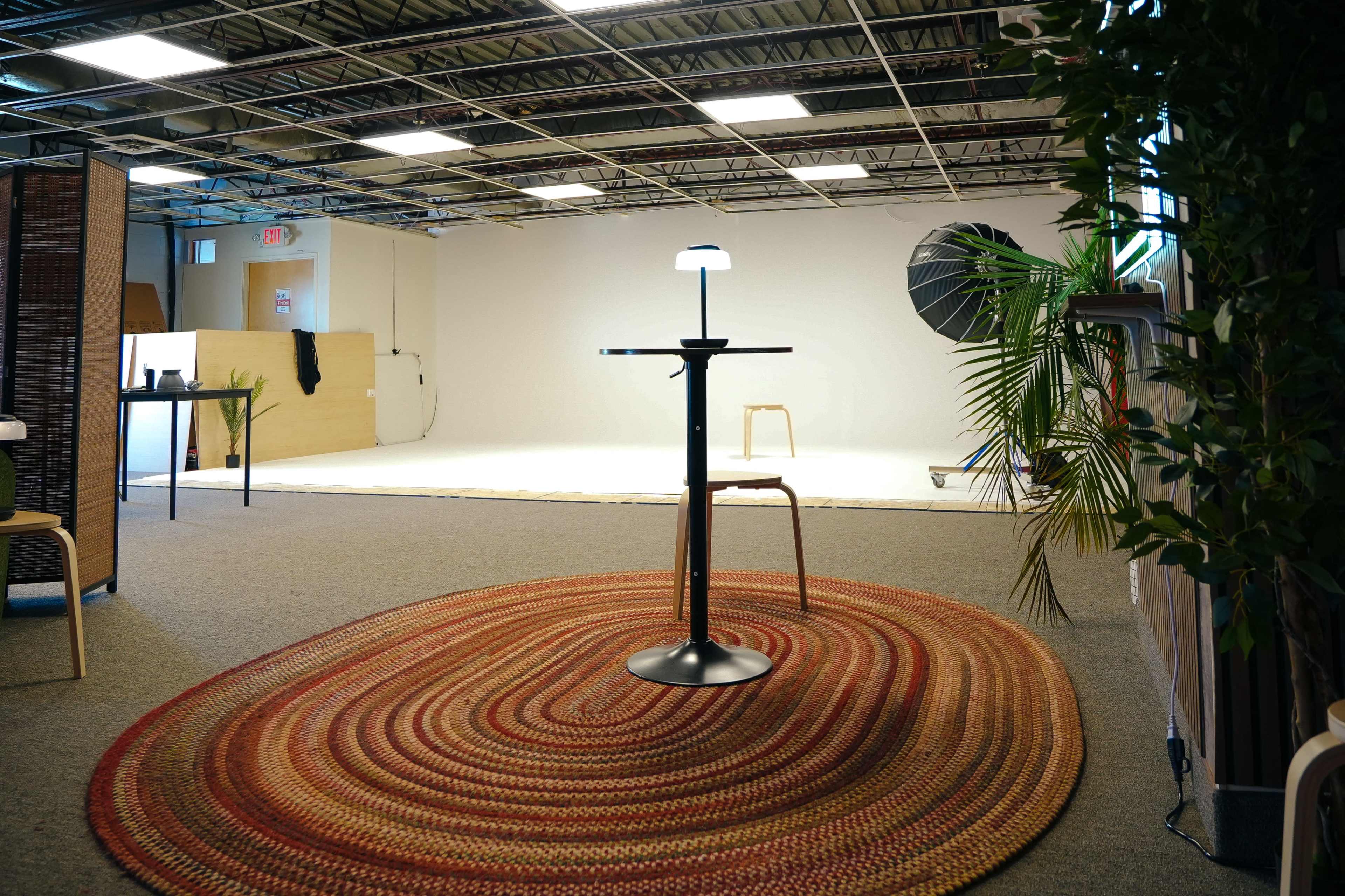 The image shows a spacious studio with a circular woven rug, a table with a lamp, and a blank backdrop in the background.