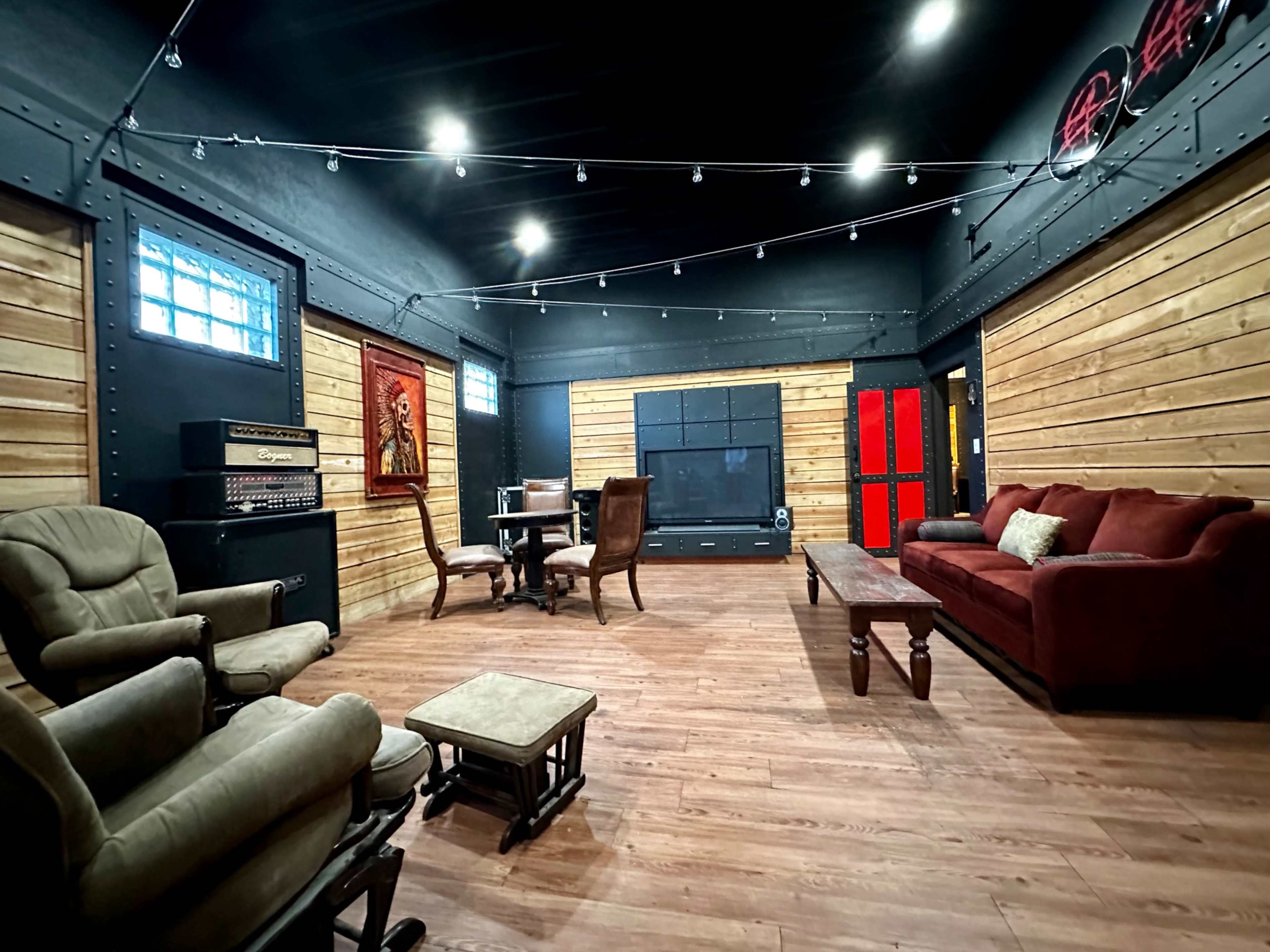The interior of a spacious room features wooden paneling, a red sofa, a dark television area, and a small dining table with chairs, all under string lights.