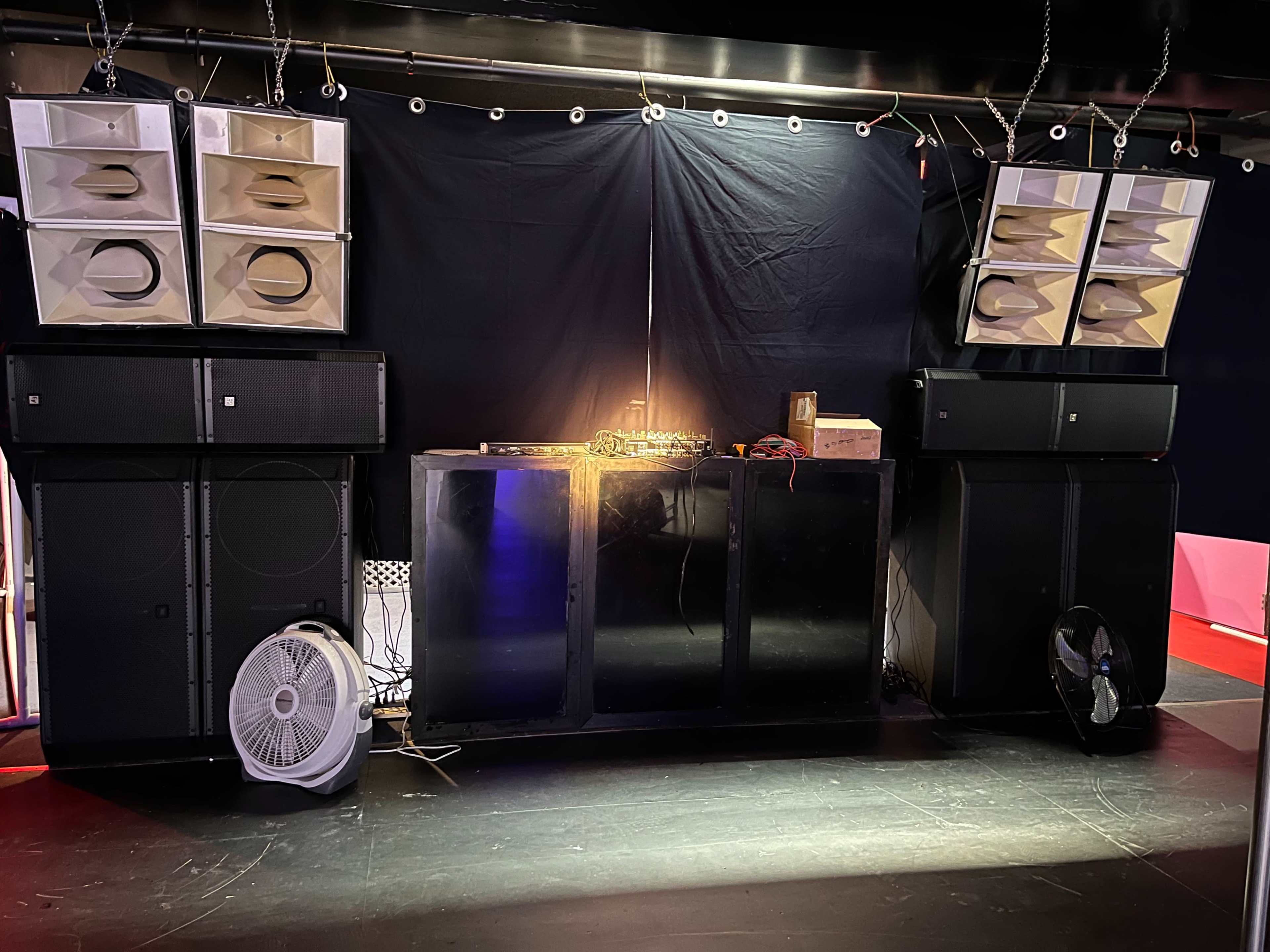 A DJ setup with large speakers on either side, a mixing console in the center, and a fan positioned on the floor.
